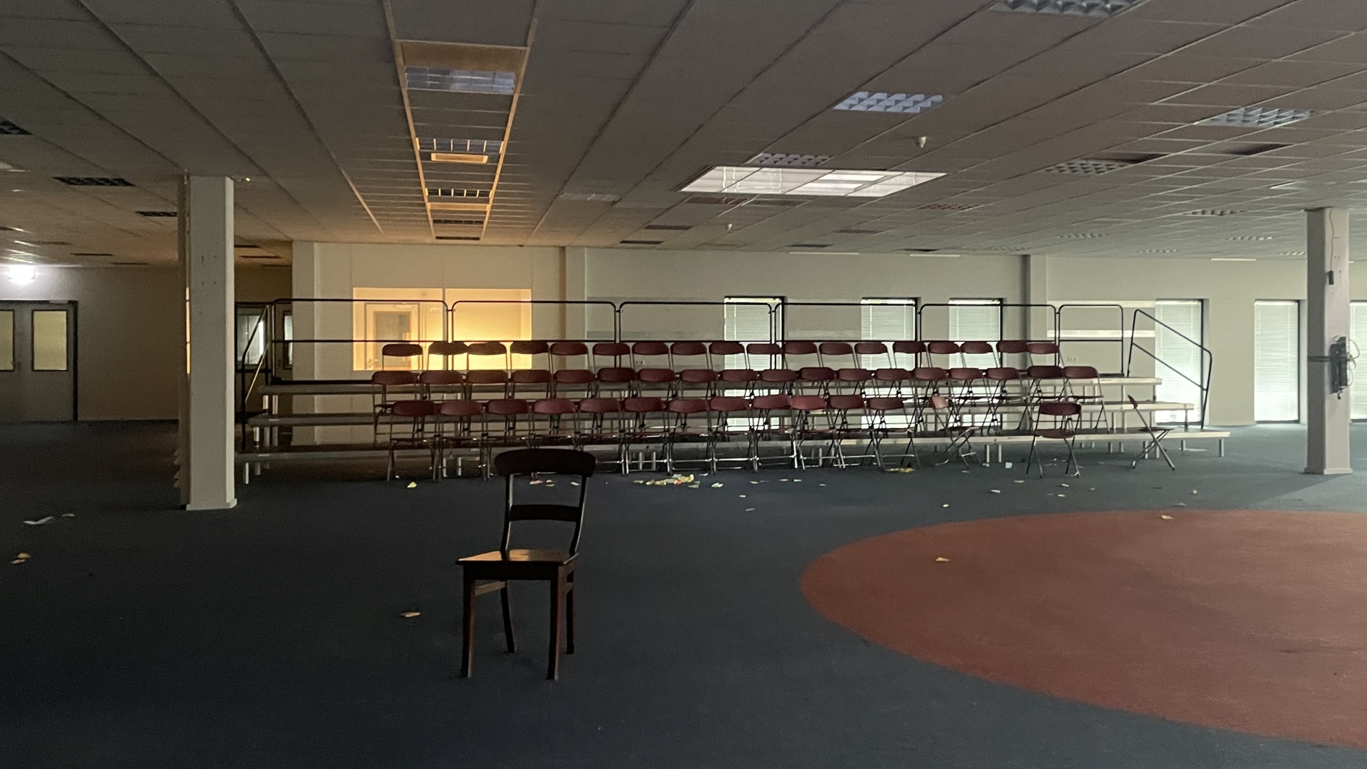 Een lege, verlichte ruimte met een tribune vol lege rode stoelen en een enkele houten stoel op de voorgrond. De ruimte heeft een systeemplafond en tl-verlichting, en er ligt afval verspreid over de vloer.