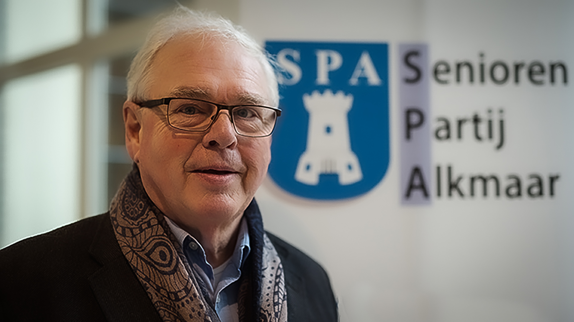 Een oudere man met bril en sjaal staat voor een bord met het logo van de Senioren Partij Alkmaar (SPA).