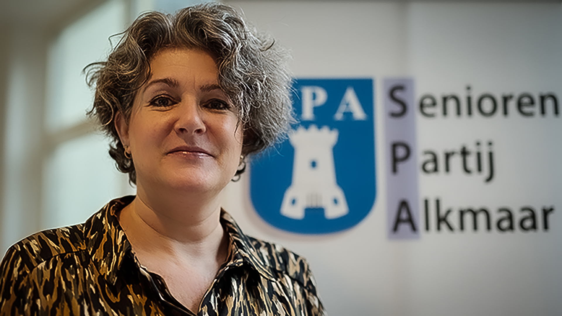 Vrouw met krullend haar en een blouse met dierlijk patroon staat voor een banner van de Senioren Partij Alkmaar, met een logo van een toren erin verwerkt.
