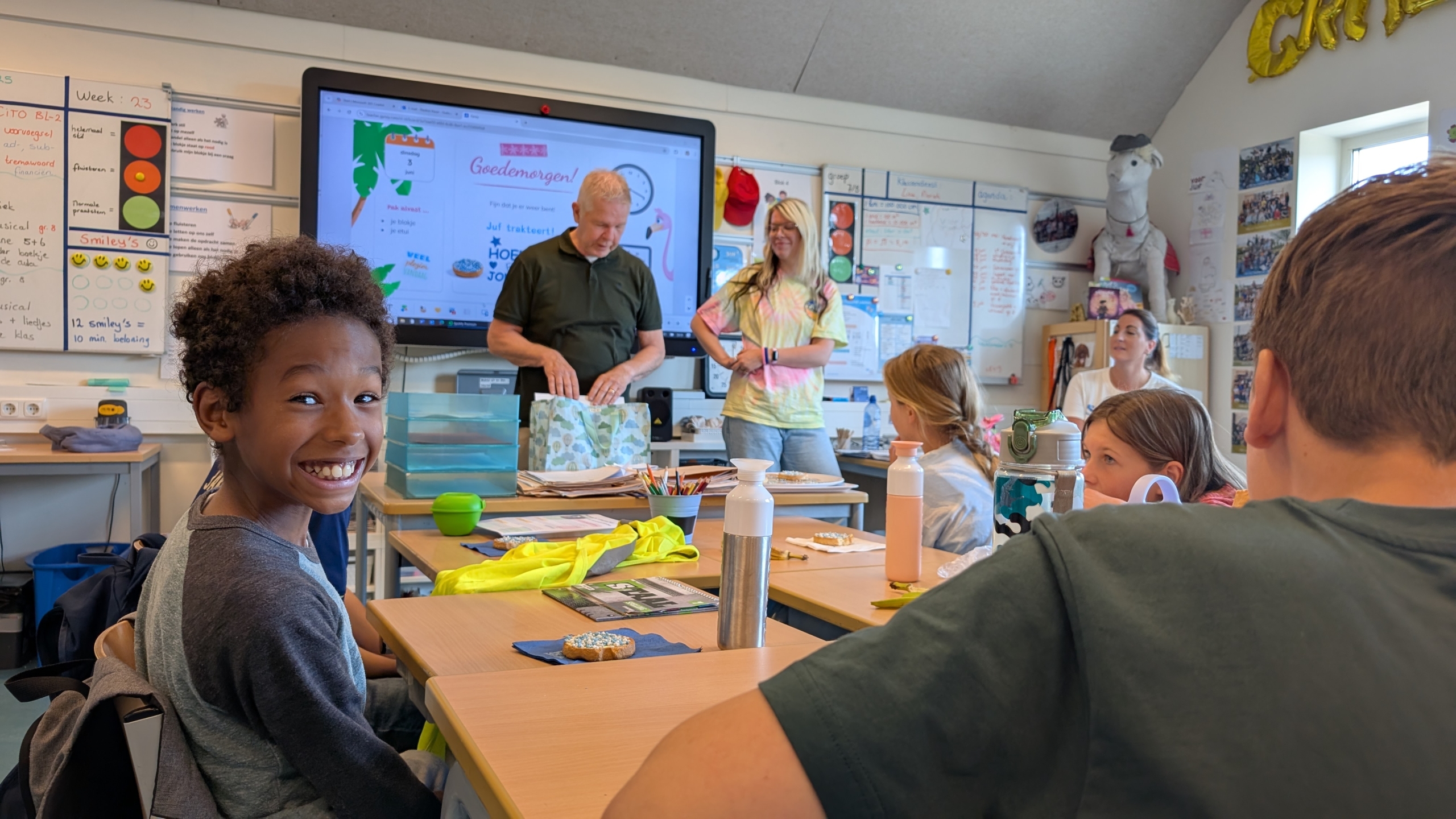Een klaslokaal met kinderen en een man en vrouw vooraan. Een scherm toont een welkomstbericht. Een jongen glimlacht enthousiast naar de camera.