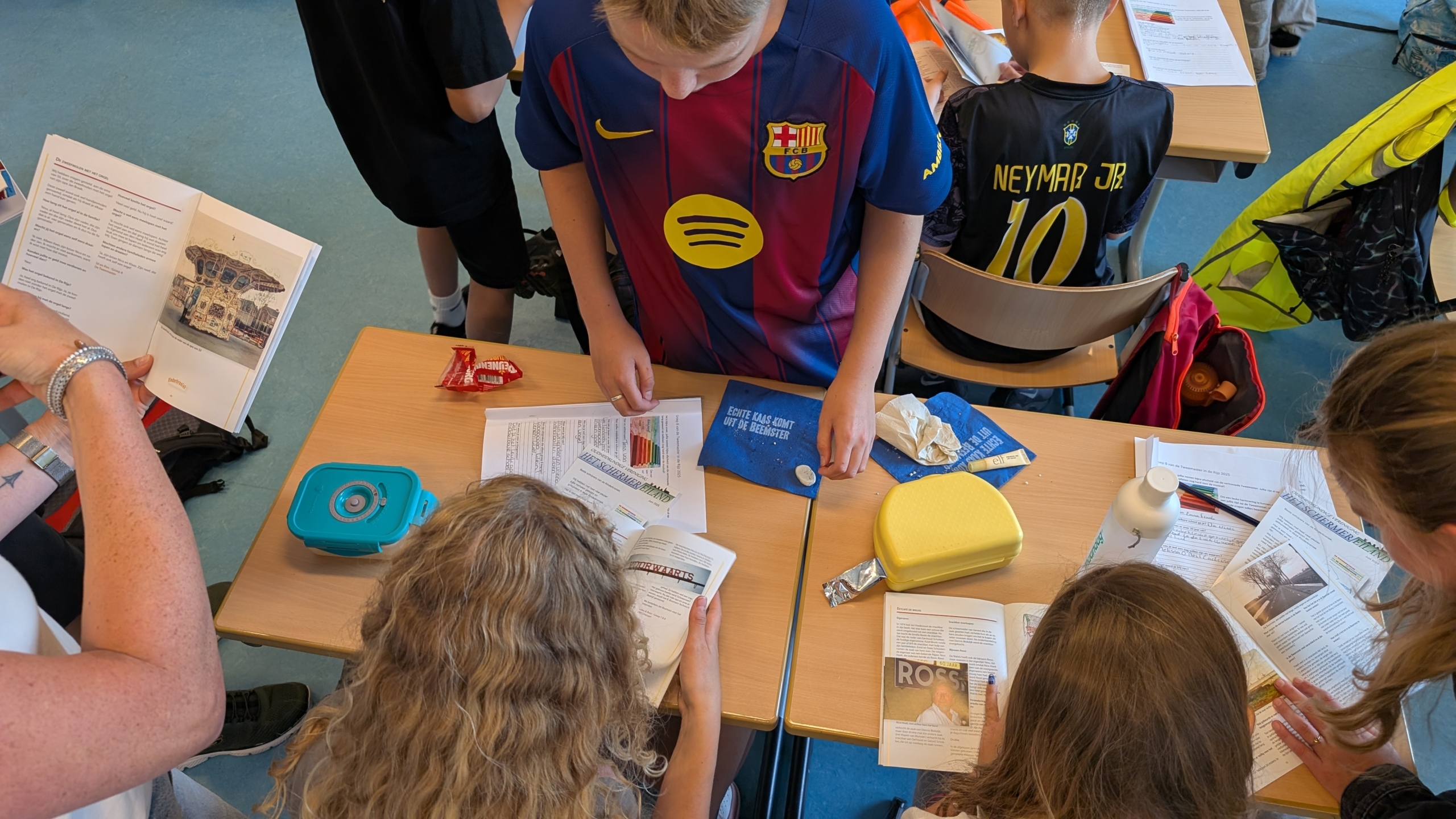 Kinderen in een klaslokaal werken samen aan tafels vol met boeken, schriften, en kleine voorwerpen. Enkele kinderen dragen sportshirts van bekende voetballers."