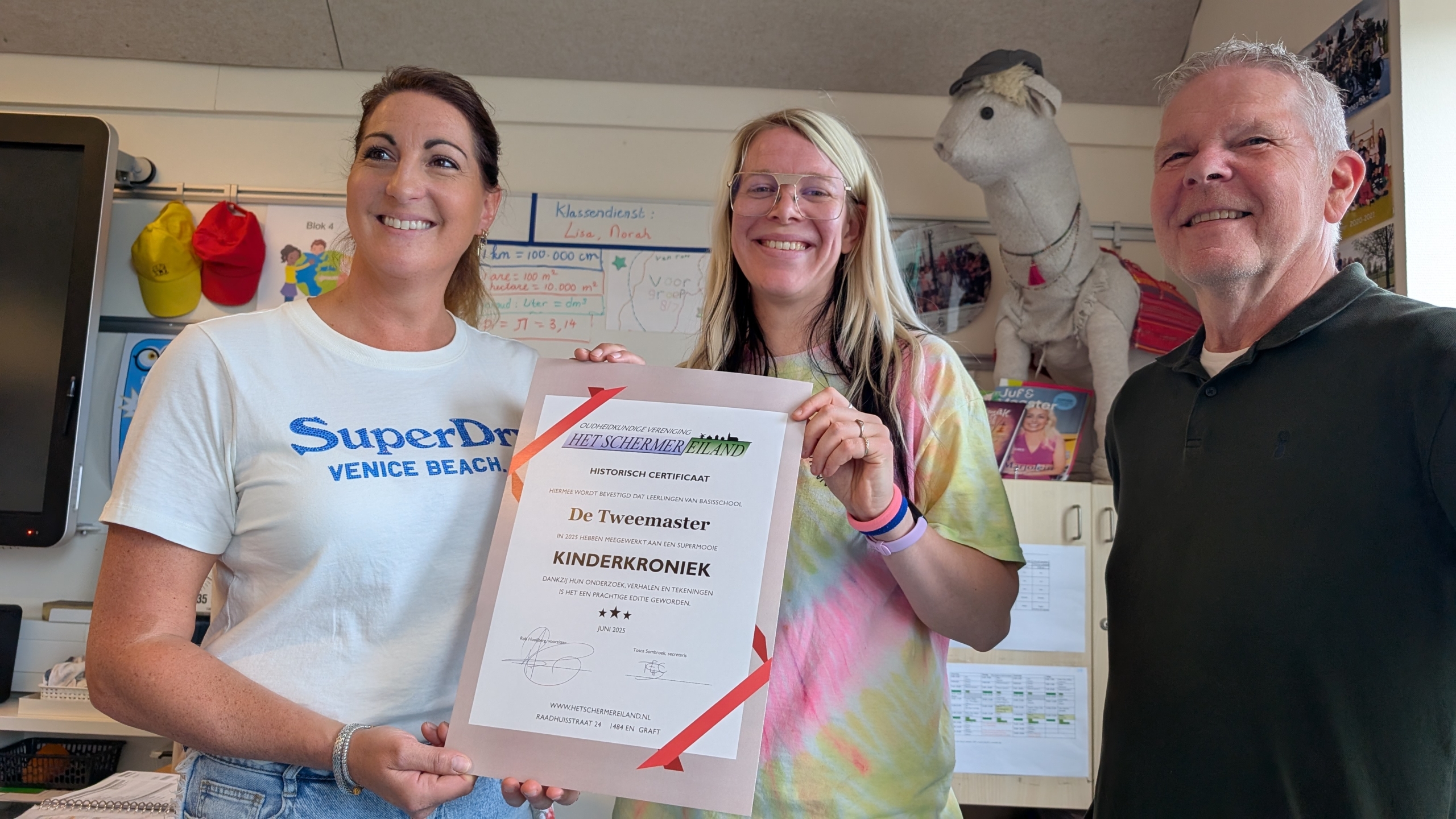 Drie mensen poseren met een certificaat in een klaslokaal, terwijl een knuffeldier op de achtergrond staat.