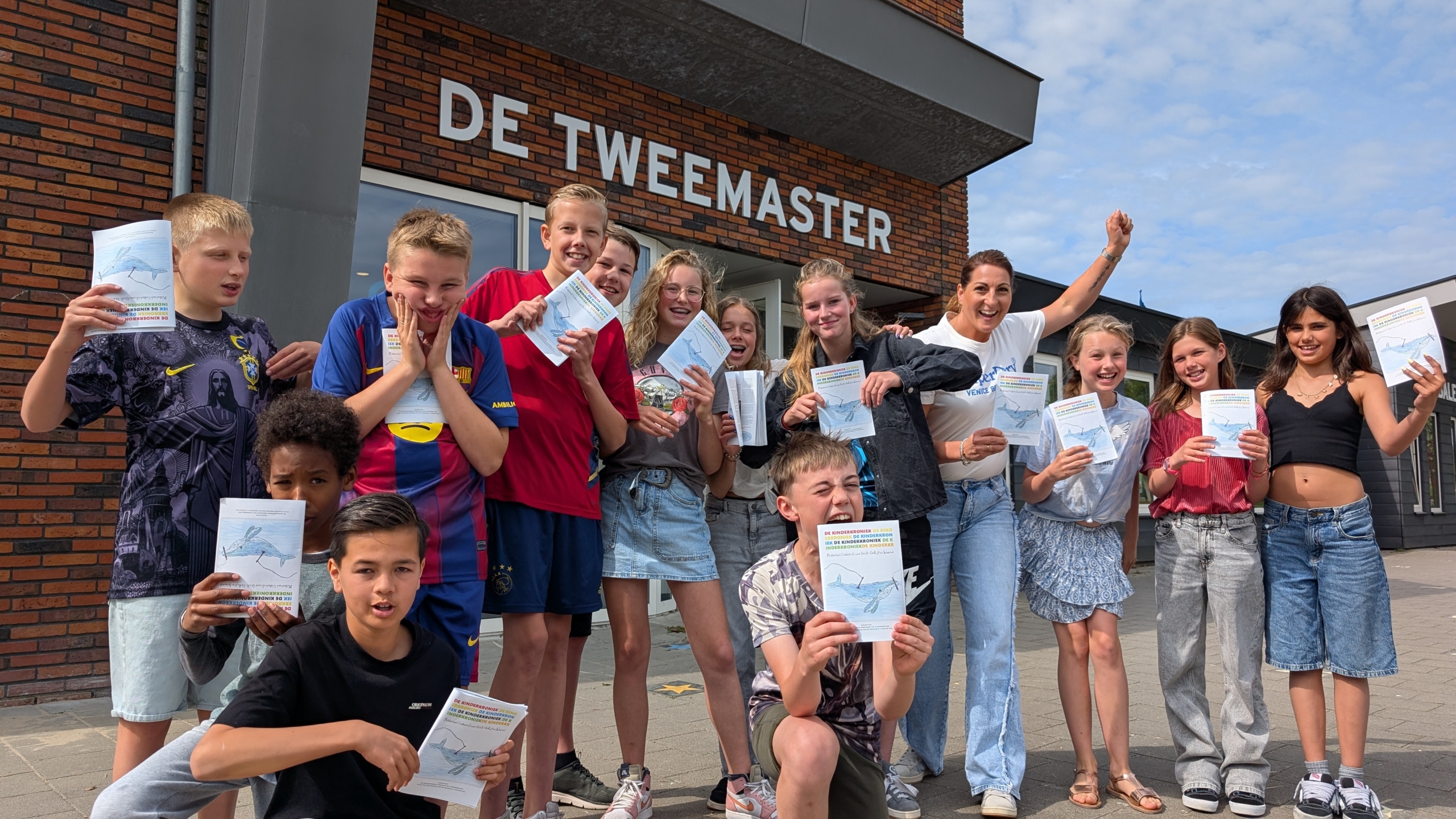 Een groep lachende kinderen en een volwassene staan buiten voor een gebouw genaamd 'De Tweemaster', terwijl ze papieren vasthouden.