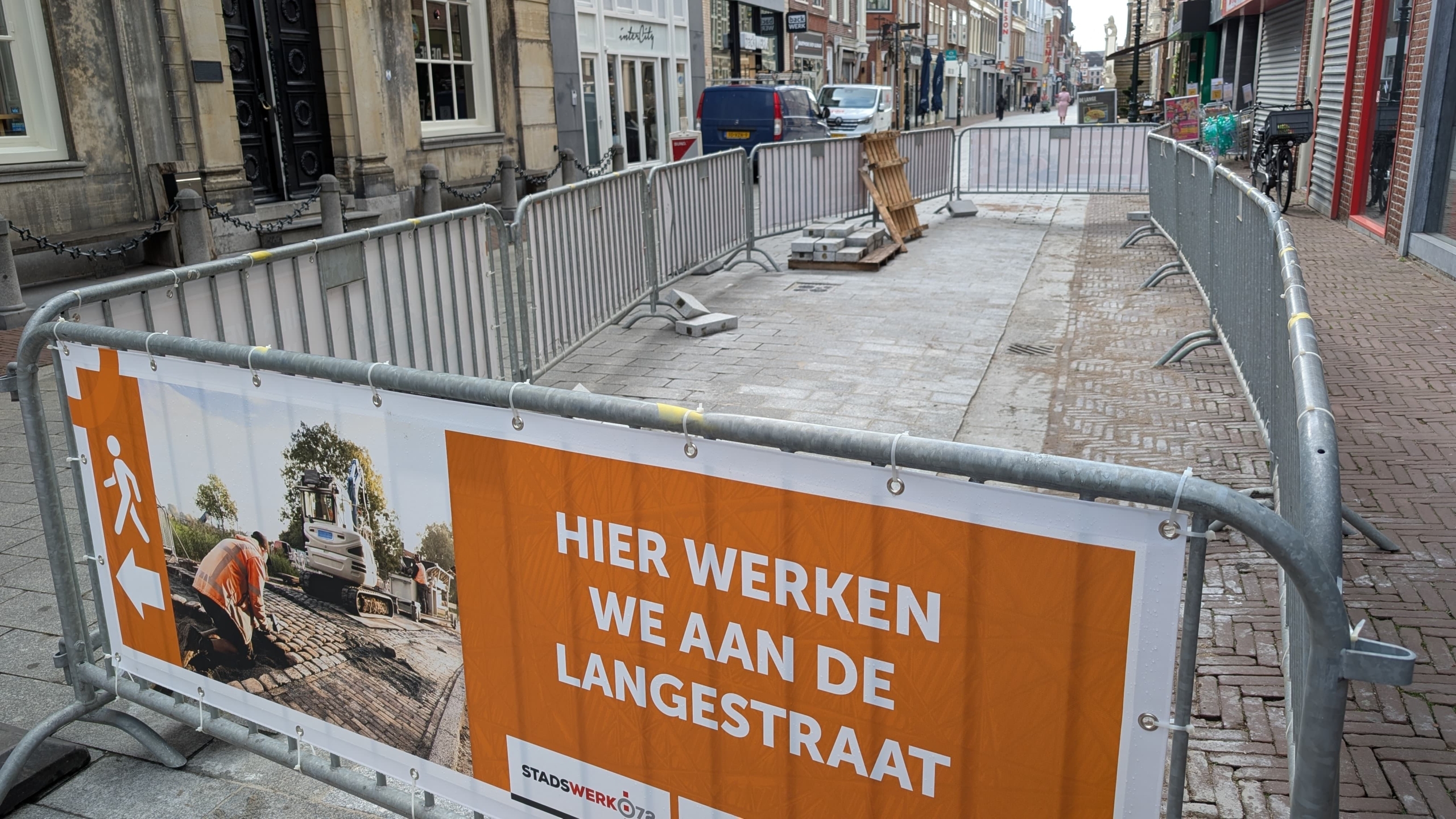 Straatwerkzaamheden in een winkelstraat, omheind met metalen hekken en een oranje bord met de tekst 'HIER WERKEN WE AAN DE LANGESTRAAT'.