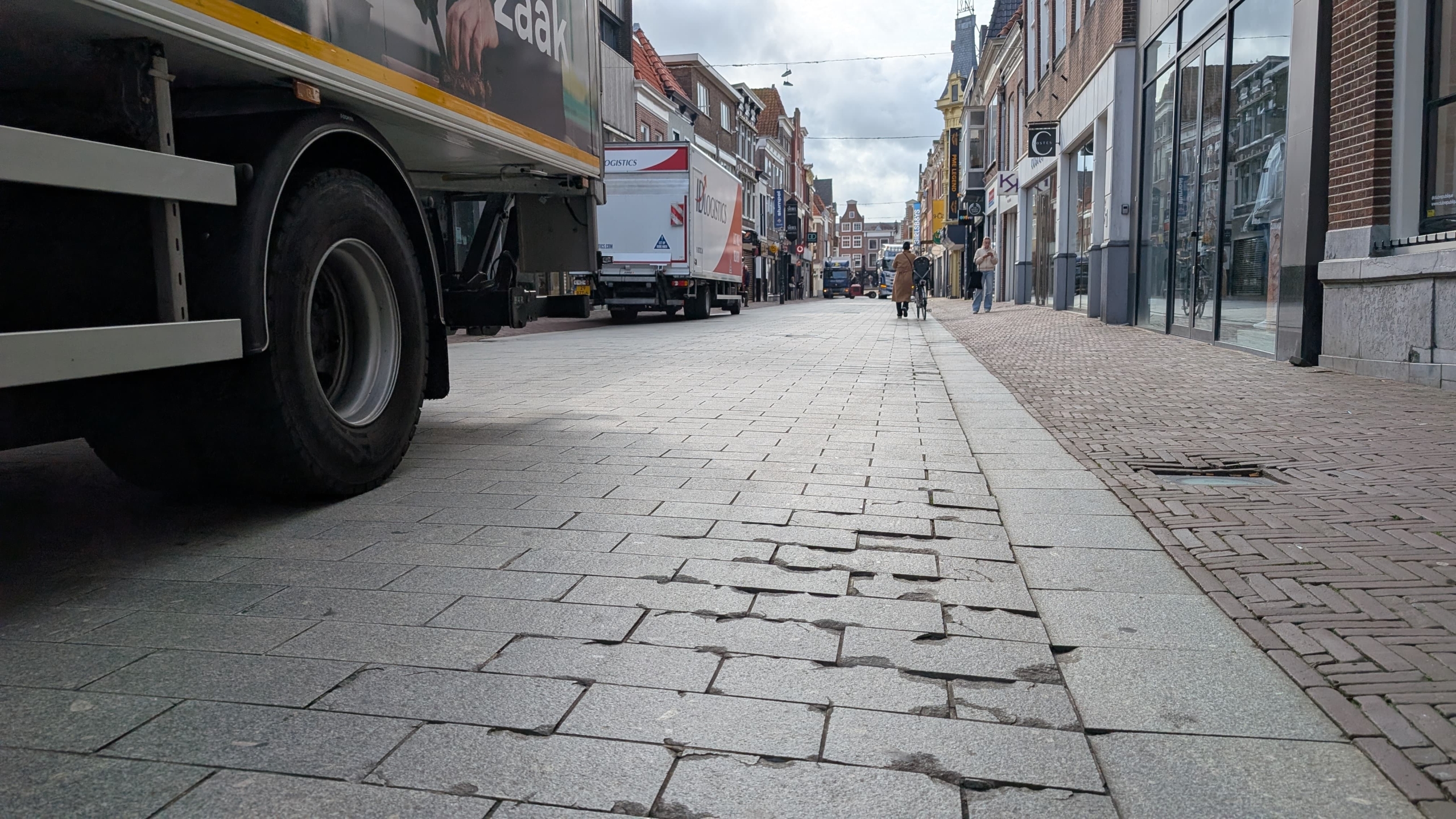 Straatbeeld van een stedelijke winkelstraat met trucks aan de zijkant geparkeerd, een stoep met beschadigde tegels, en enkele voetgangers in de verte.