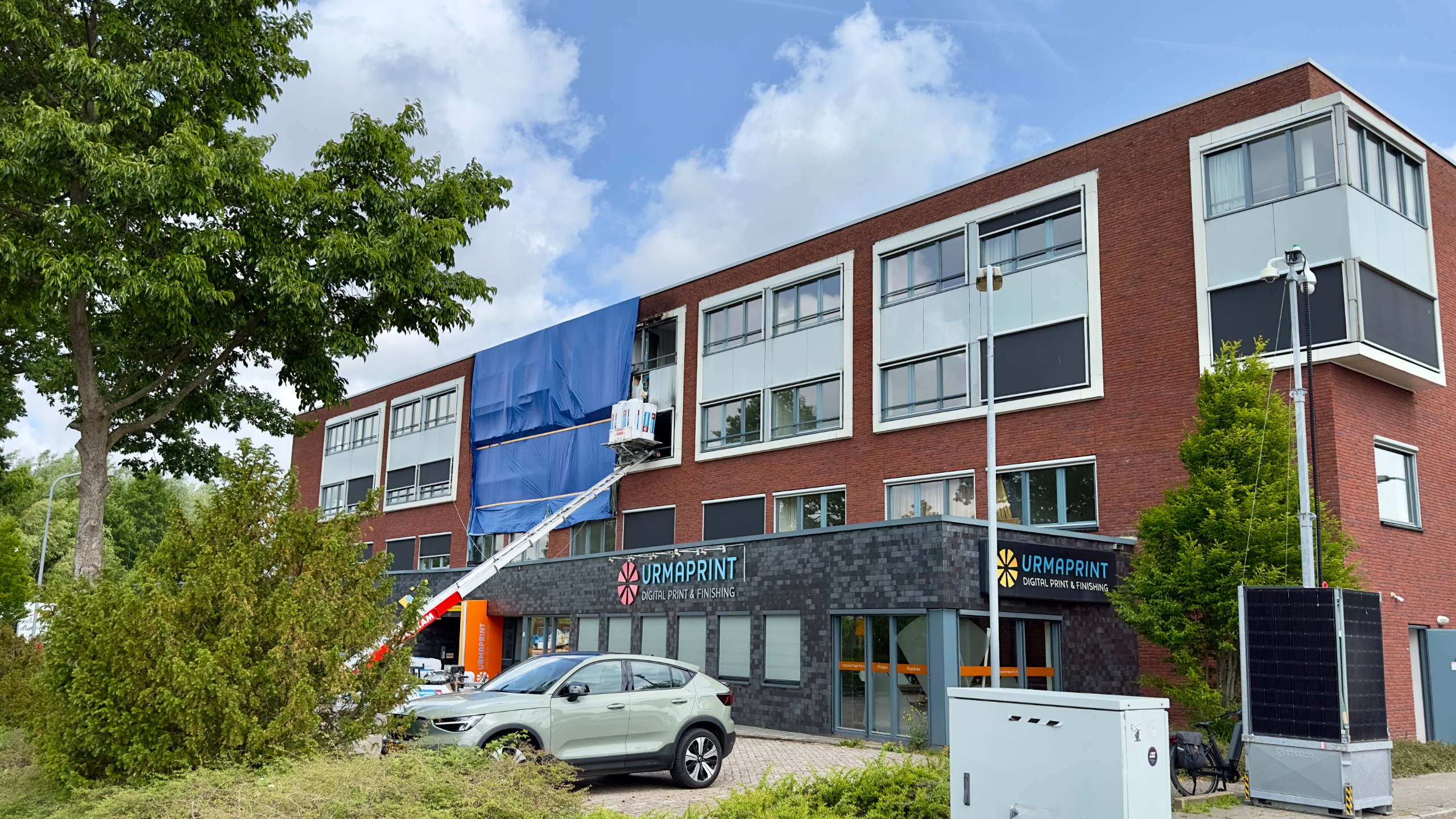Een bakstenen bedrijfsgebouw met meerdere verdiepingen, waarbij een deel bedekt is met een blauw zeil. Er staat een hoogwerker met een persoon bezig aan het gebouw. Op de begane grond is er een bedrijf genaamd 'Urmaprint'. Voor het gebouw staat een geparkeerde auto en er zijn bomen en struiken zichtbaar.