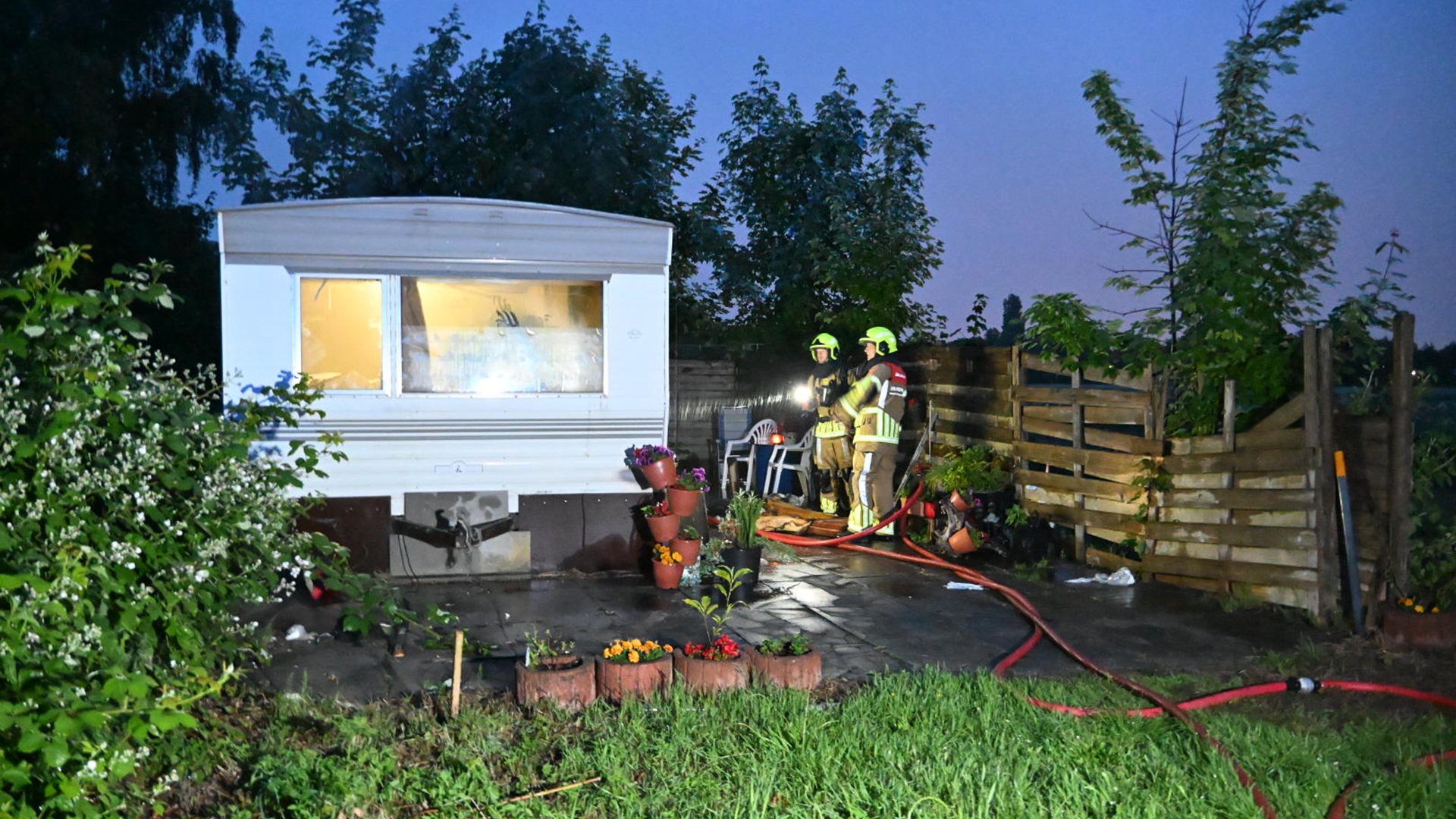 Twee brandweermannen in gele helmen staan naast een witte caravan in een tuin met planten en bloemen, in de schemering.