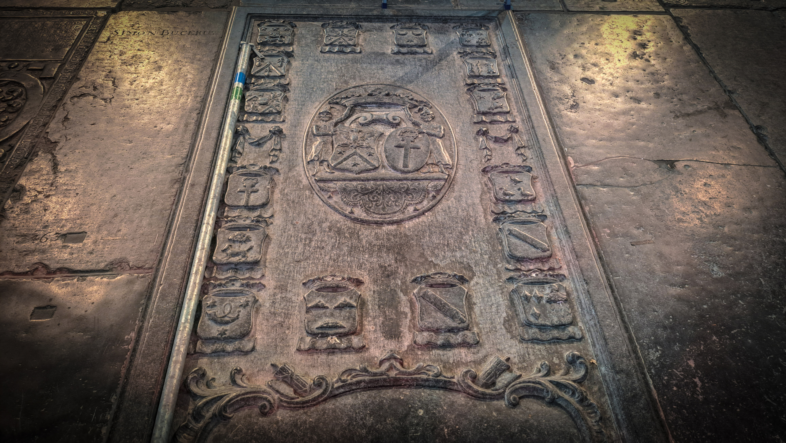 Gedenksteen met gegraveerde wapenschilden en een ovaal ontwerp in het midden, met een kruis en decoratieve elementen, omringd door verschillende kleinere symbolen op een metalen oppervlak.