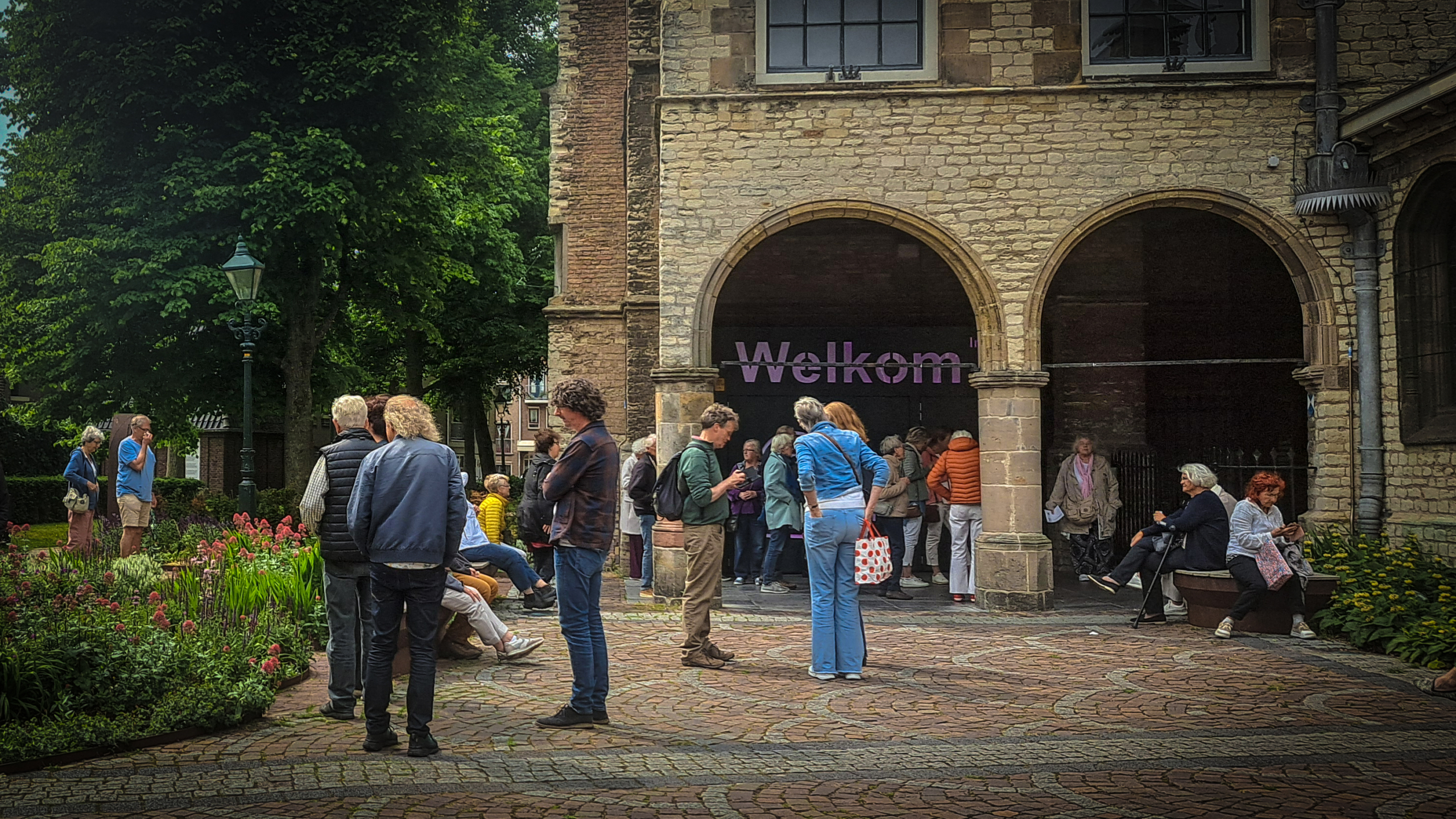 Groep mensen staat en zit in een tuin voor een steenachtig gebouw met een boogvormige ingang waarop 'Welkom' staat geschreven. Er zijn groene bomen en bloeiende bloemen op de voorgrond.