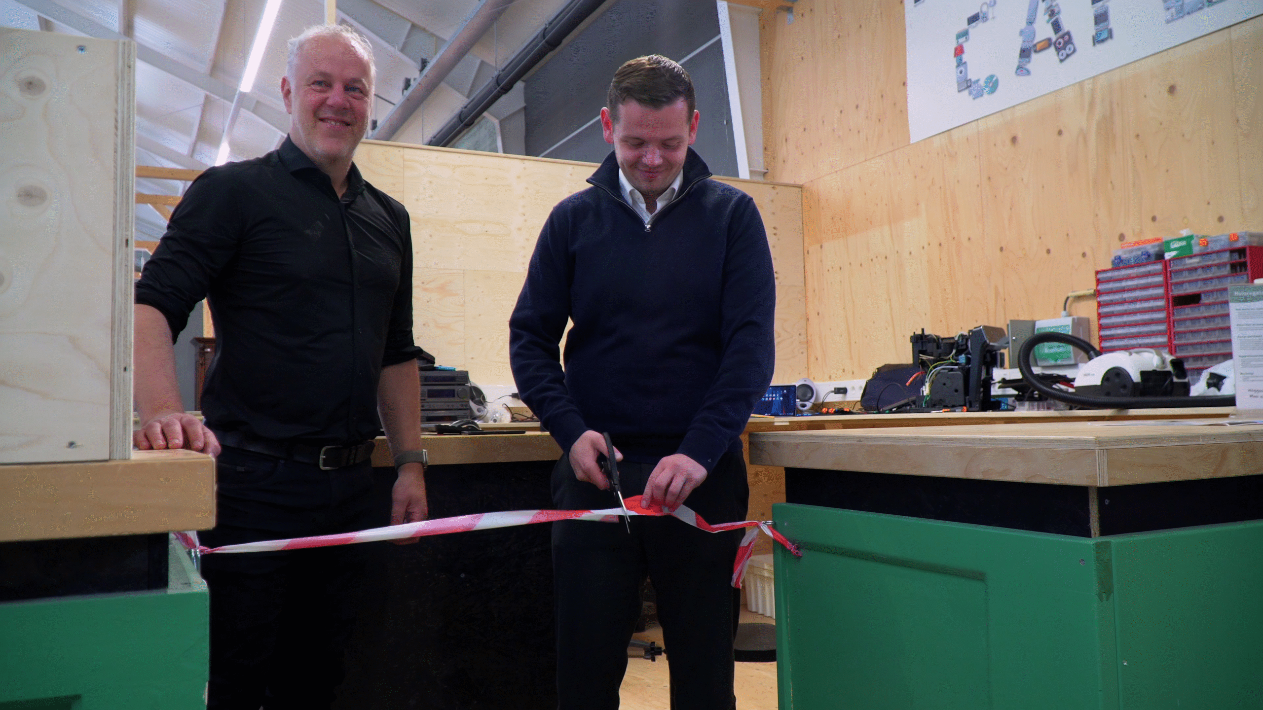 Twee mannen zijn in een werkplaats met houten wanden en apparatuur op de achtergrond. De man rechts knipt een rood-wit lint door met een schaar terwijl de andere man naast hem staat en glimlacht.