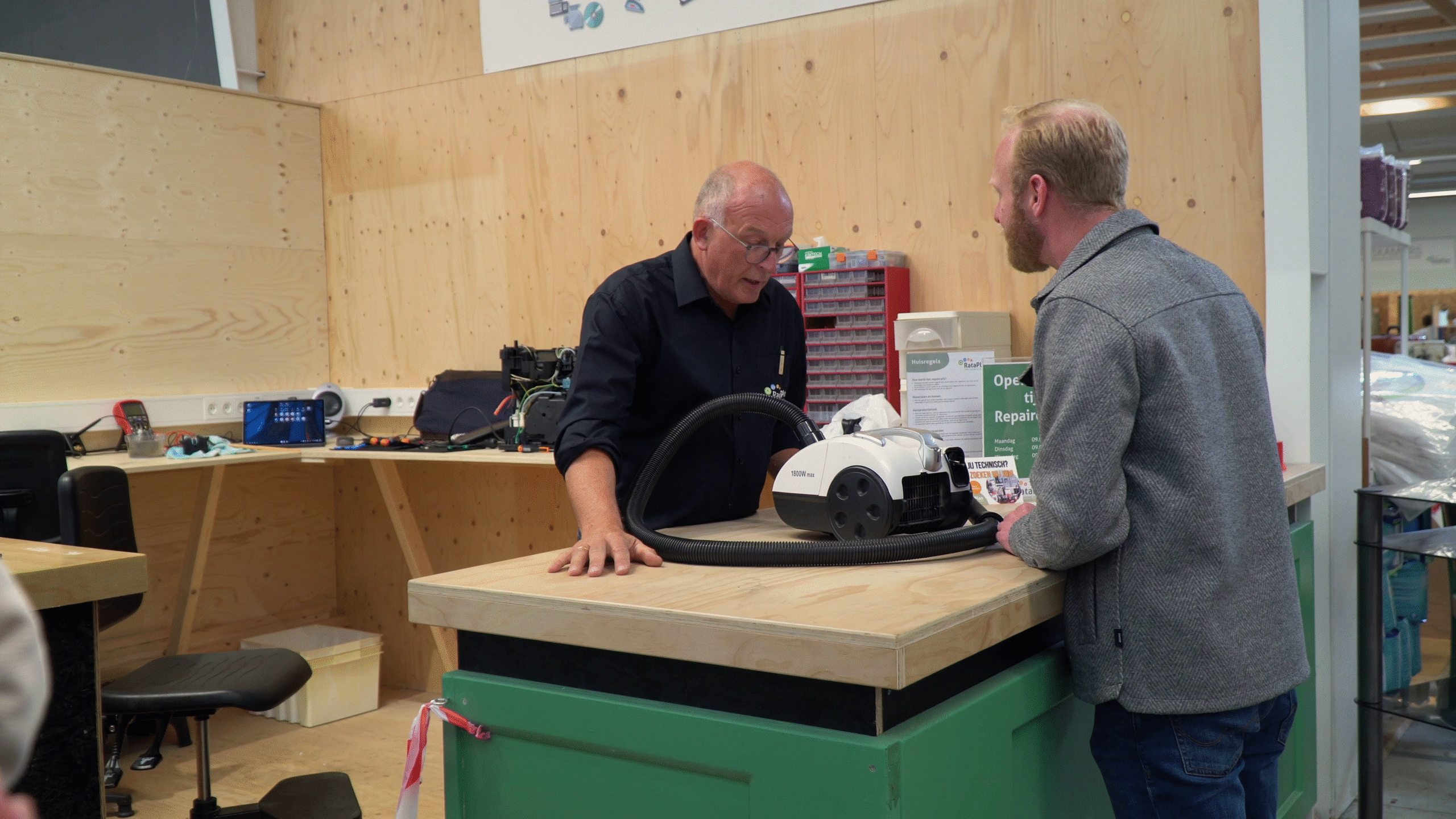 Twee mannen in gesprek over een stofzuiger in een werkplaats met houten muren en een werkbank vol gereedschap.