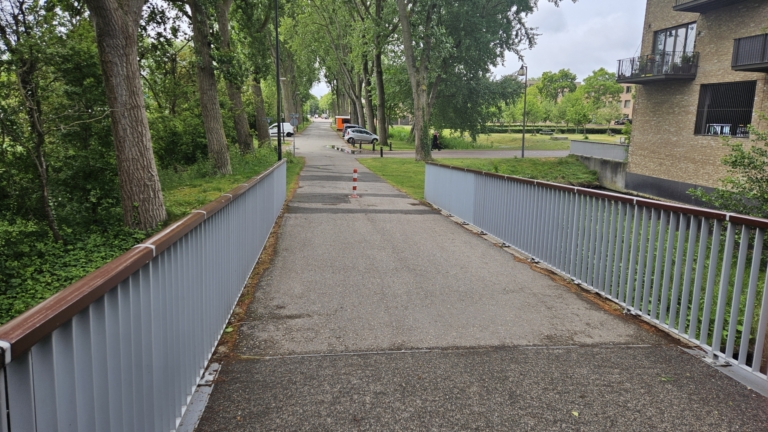 Een smalle brug met leuningen loopt over een sloot richting een pad dat langs hoge bomen en een bakstenen woongebouw met balkons voert.
