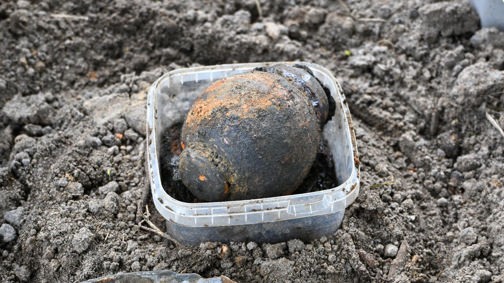 Een roestige granaat ligt in een plastic bakje op de grond, omgeven door aarde.