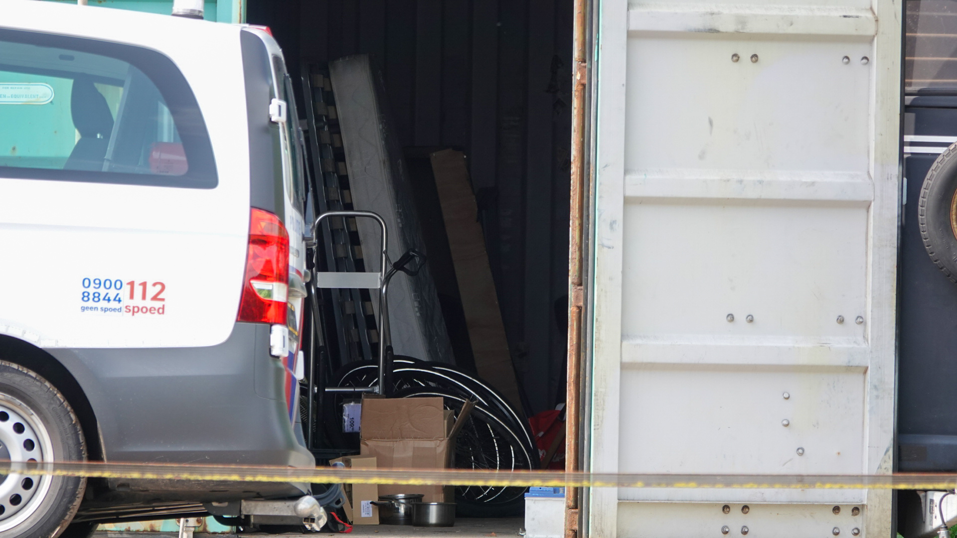 Een politievoertuig naast een openstaande container met binnenin onder andere kartonnen dozen en metalen voorwerpen.