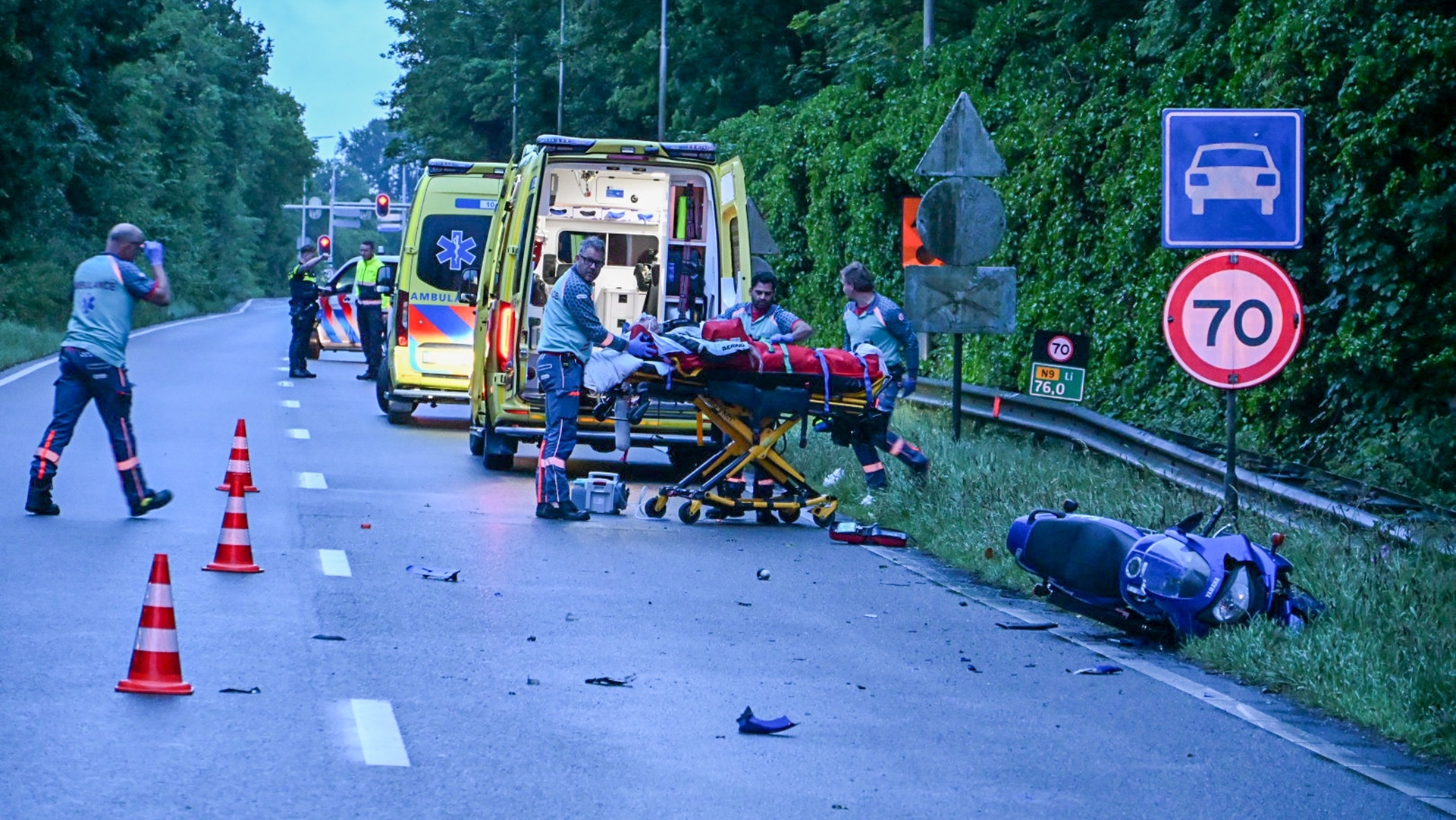 Hulpverleners bij een open ambulance behandelen een slachtoffer op een brancard na een verkeersongeval, met een omgevallen scooter op de weg en snelheidsborden op de achtergrond.