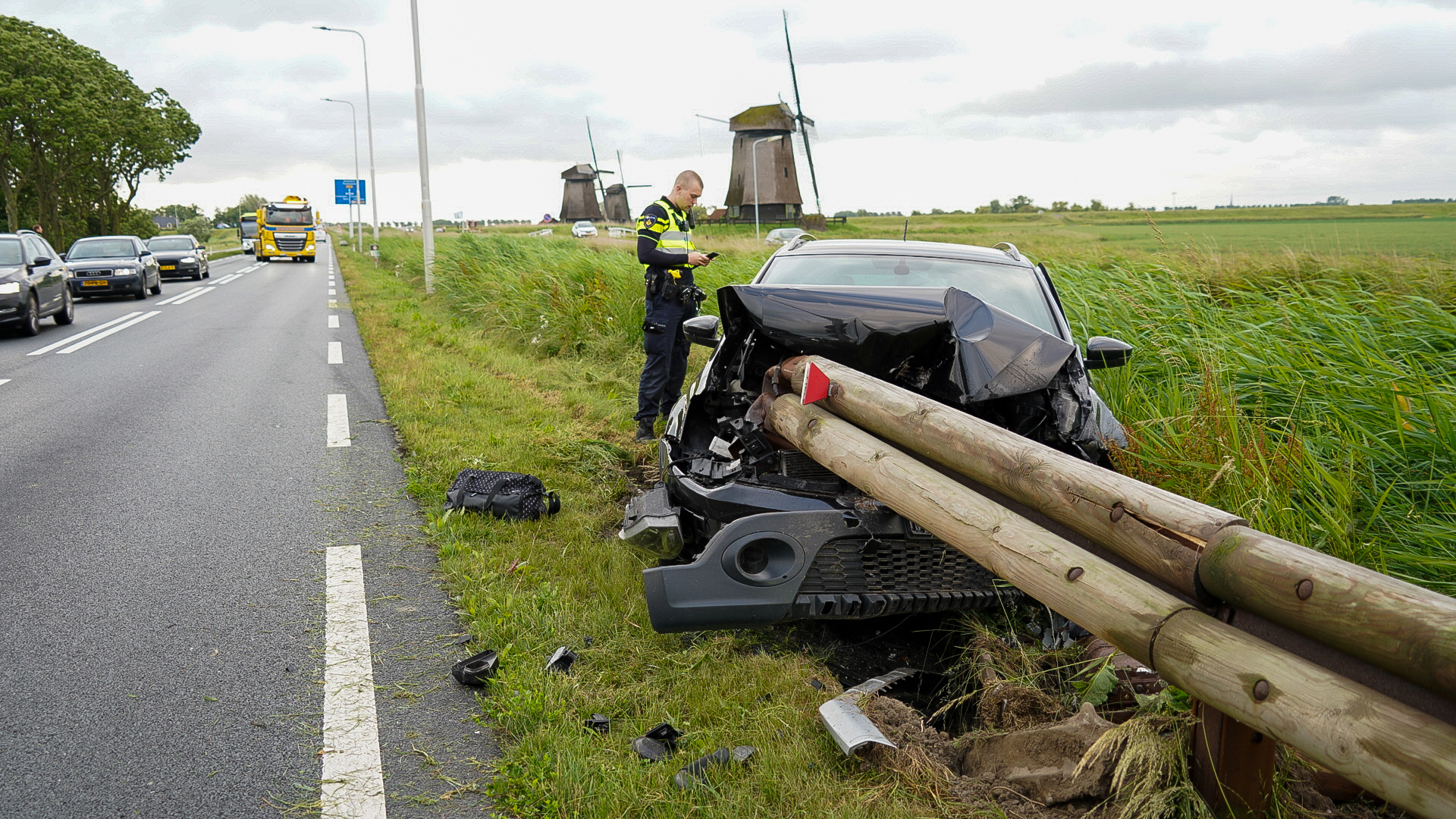 Autowrak na een ongeluk met een vangrail langs een weg, met een agent die notities maakt en windmolens op de achtergrond.
