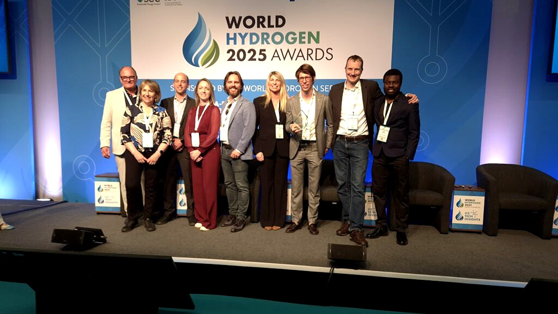Een groep mensen poseert voor een foto bij een podium tijdens de World Hydrogen 2025 Awards, genomen in een conferentieomgeving met een groot bord met het evenementlogo op de achtergrond.