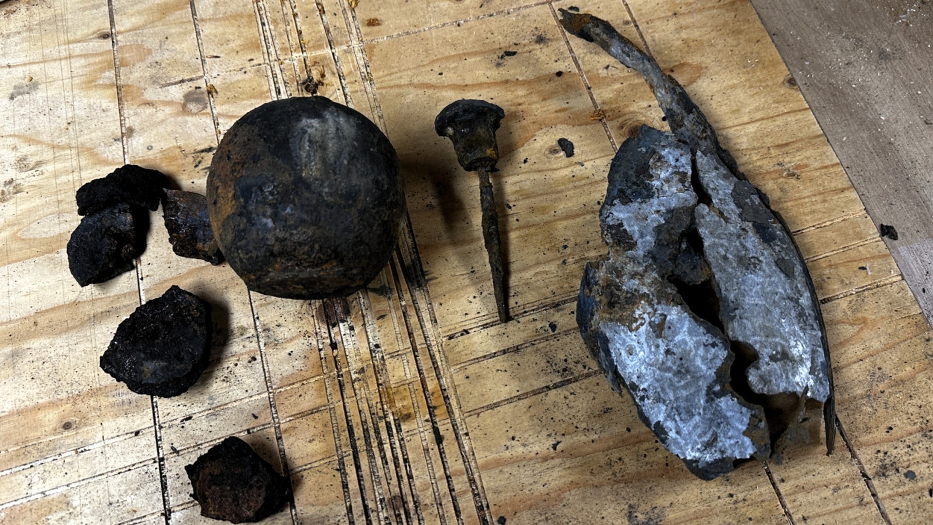 Diverse roestige metalen voorwerpen, waaronder een bol, spijker en gebroken plaat, liggen op een houten oppervlak.