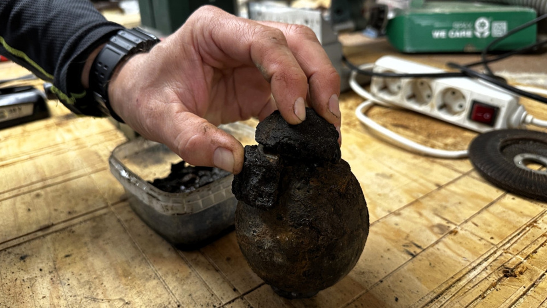 Een roestige, bolvormige kogel wordt vastgehouden door een hand, terwijl er brokken zwart materiaal op de tafel liggen, omringd door gereedschappen en een stopcontactdoos in een werkplaatsomgeving.