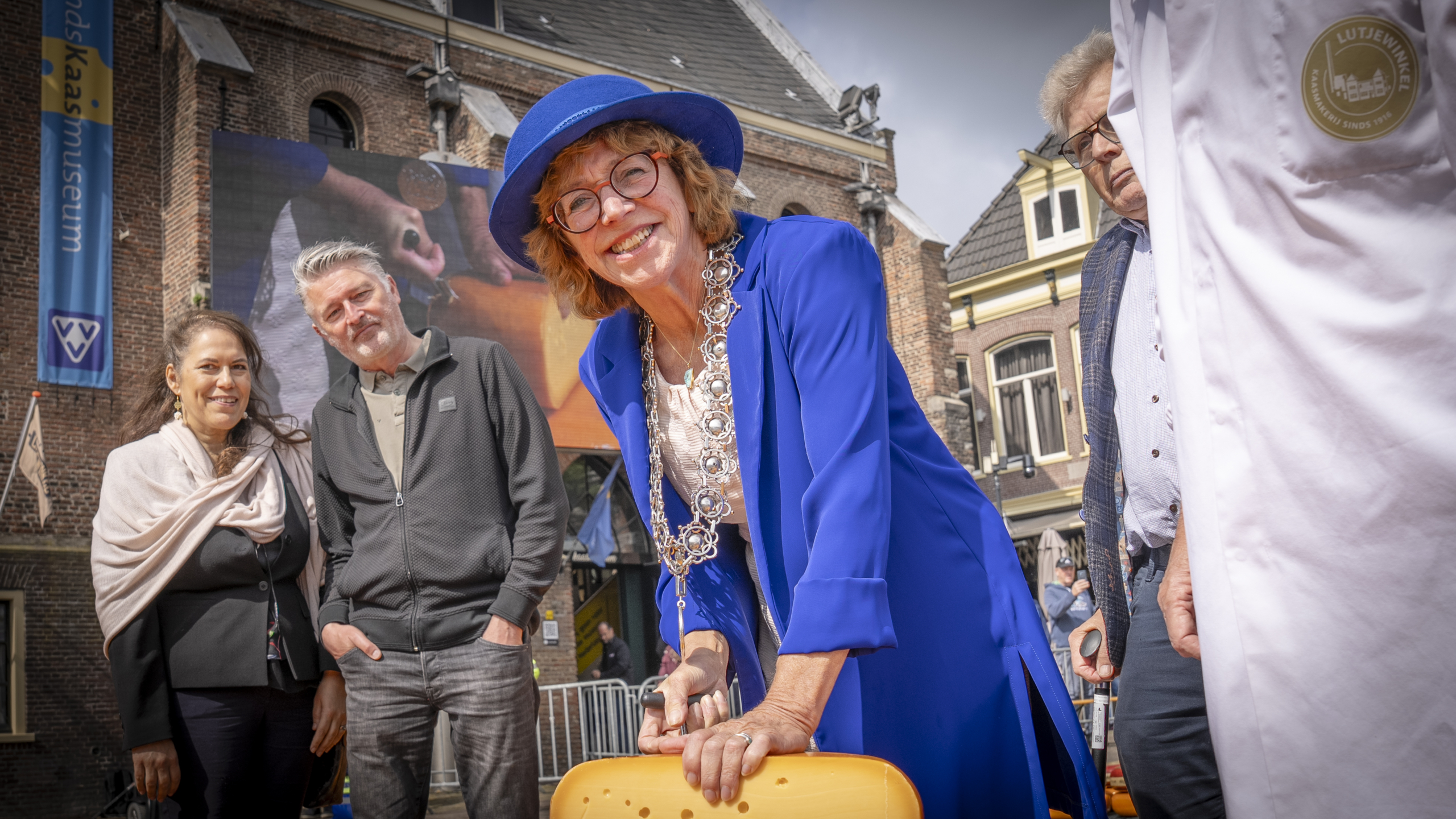 Een vrouw in een blauwe hoed en mantel lacht terwijl ze met een kaas werkt op een plein voor een historisch gebouw, omringd door een groep mensen.