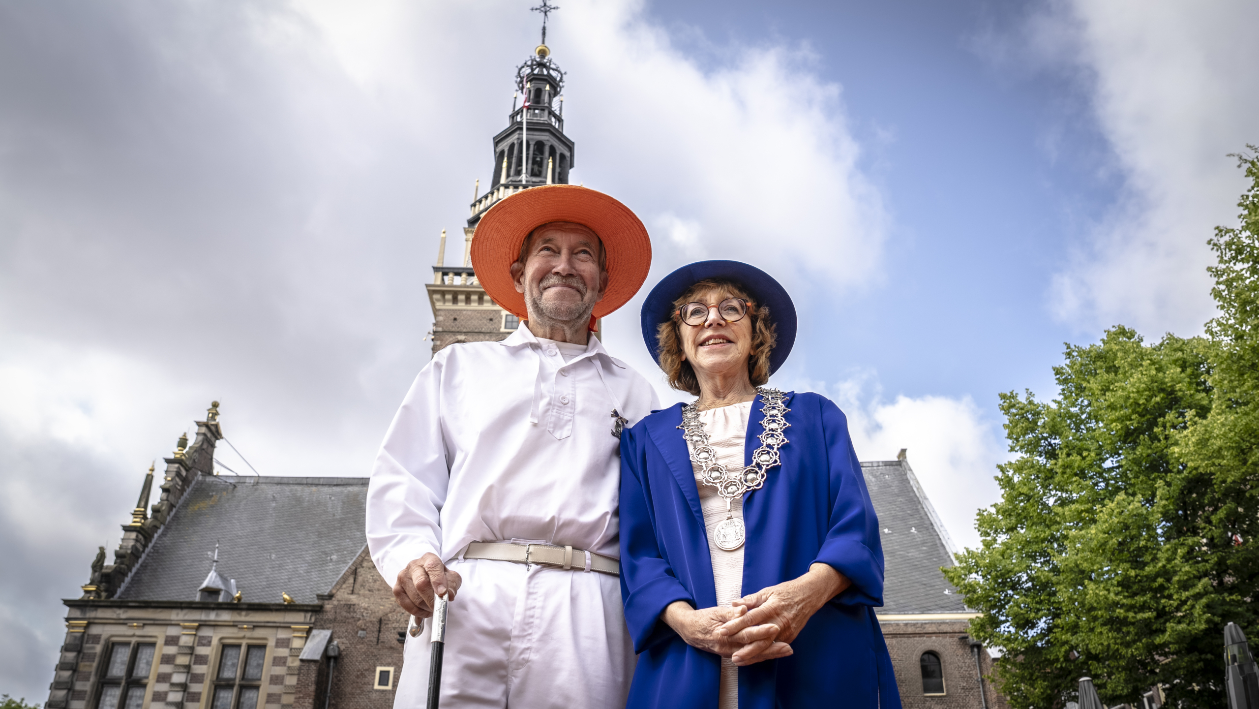 Twee oudere personen in kleurrijke kleding staan voor een historische toren, de man in het wit met een oranje hoed en wandelstok, en de vrouw in een blauwe mantel met ketting en hoed.