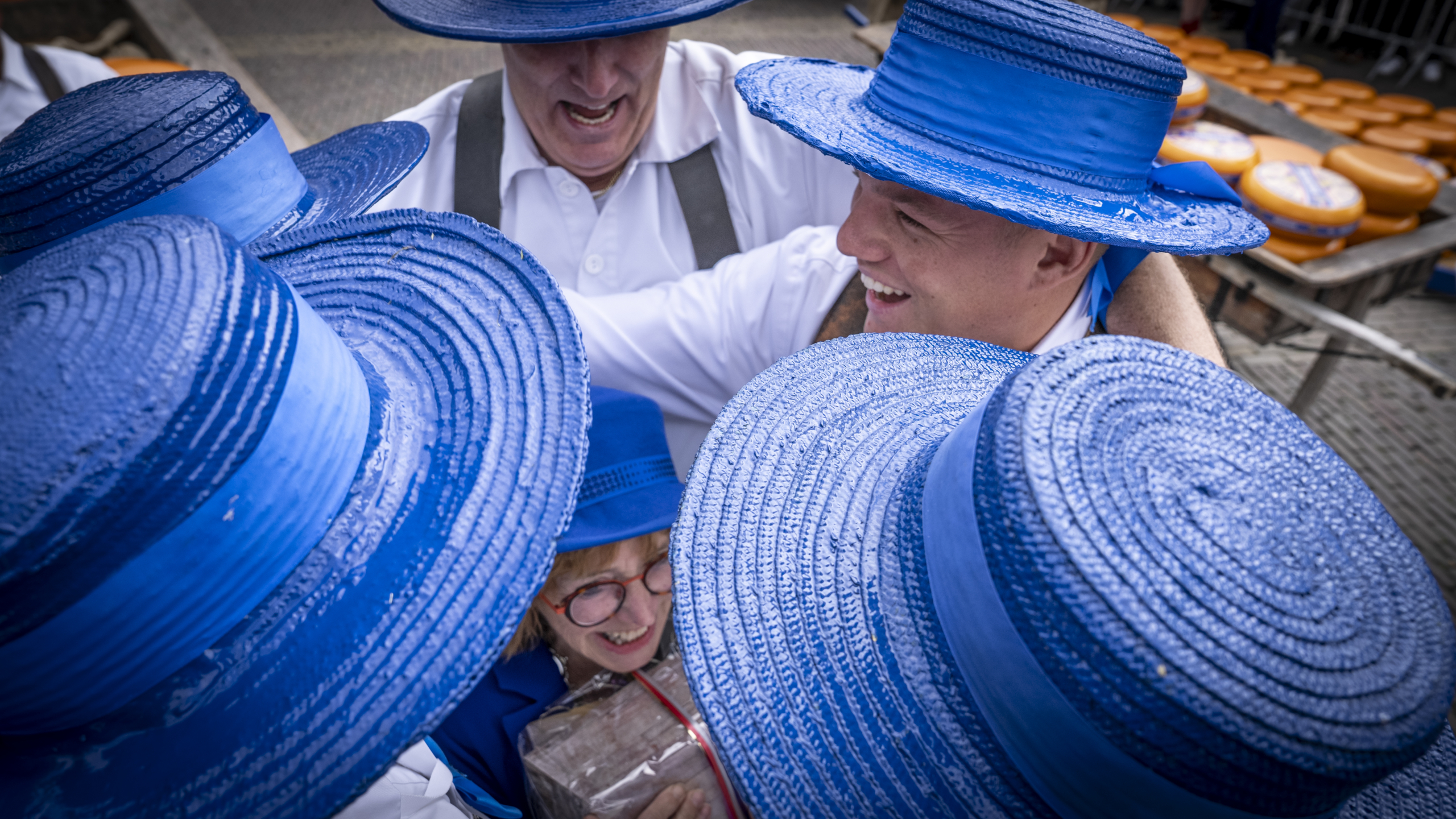 Mensen dragen blauwe hoeden en witte kleding, lachen en omhelzen elkaar op een festival, met in de achtergrond stapels kaas.