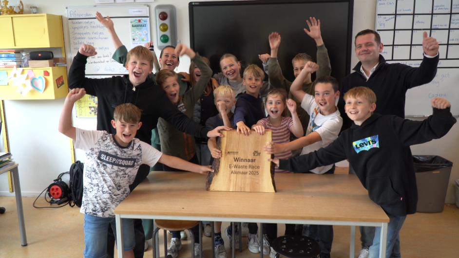 Een groep blije kinderen en een volwassen man poseren in een klaslokaal met een trofee waarop "Winnaar E-Waste Race Alkmaar 2025" staat.