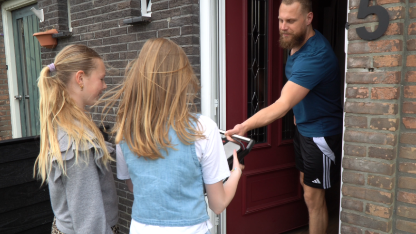 Twee meisjes staan bij een rode deur waar een man met een baard hen iets aanbiedt. Hij draagt een blauw shirt en korte zwarte broek. Ze staan voor een huis met een bakstenen muur.