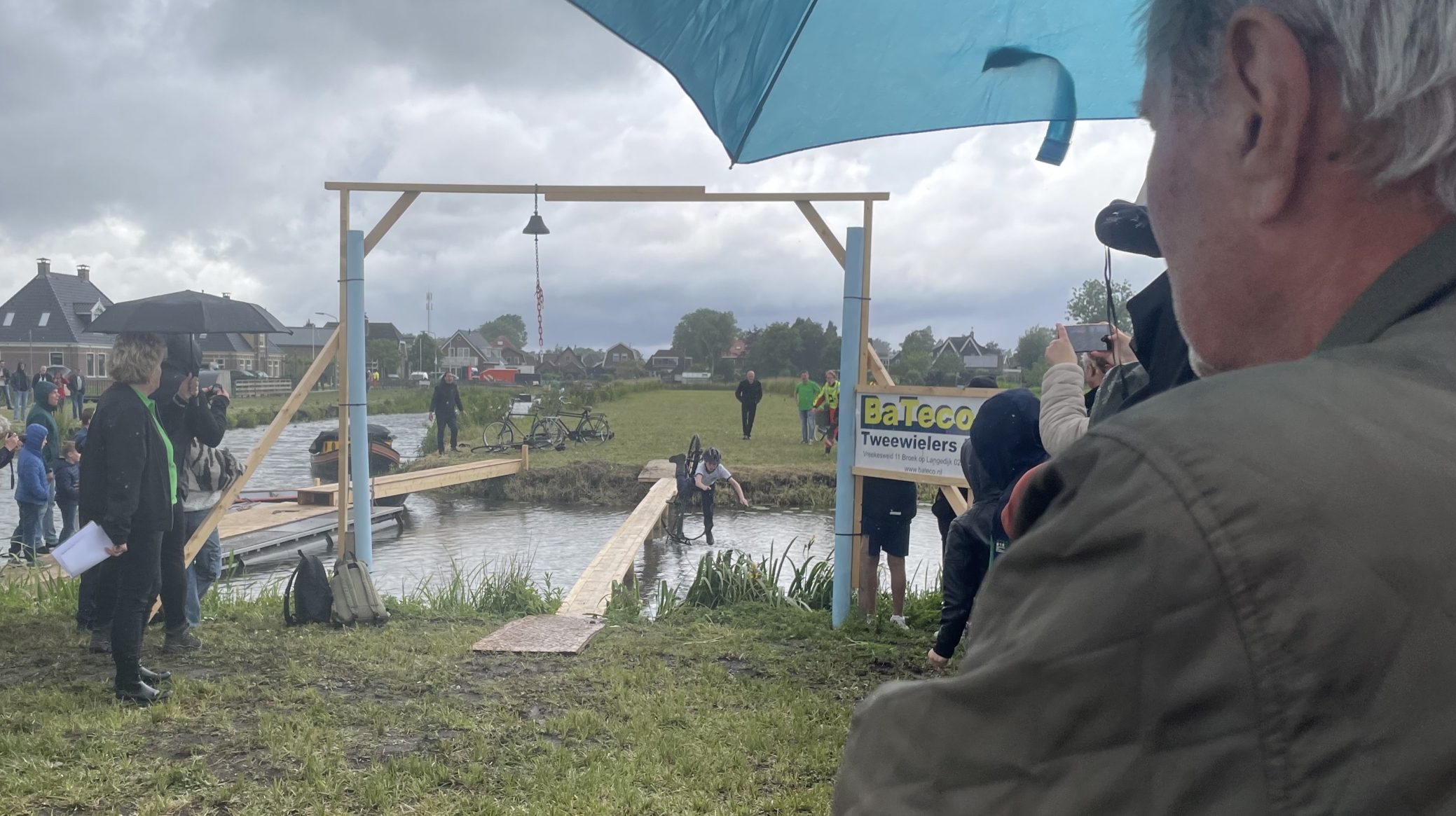 Mensen bekijken een evenement in een regenachtige omgeving met een houten constructie boven een smalle brug over het water. Een deelnemer met een fiets lijkt in het water te vallen terwijl omstanders foto's maken en paraplu's gebruiken.