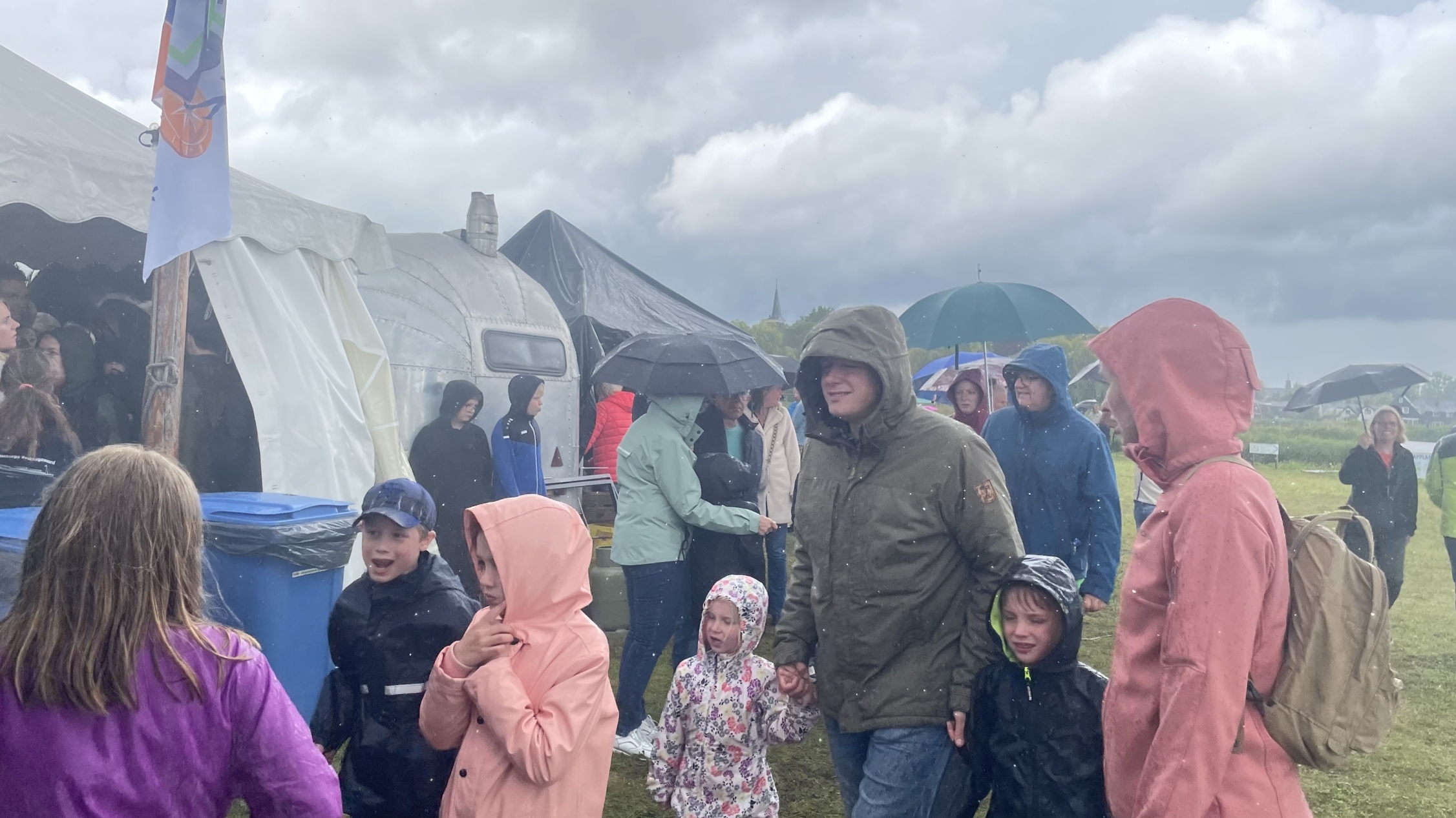 Mensen in regenkleding lopen buiten bij een evenement onder een bewolkte hemel.