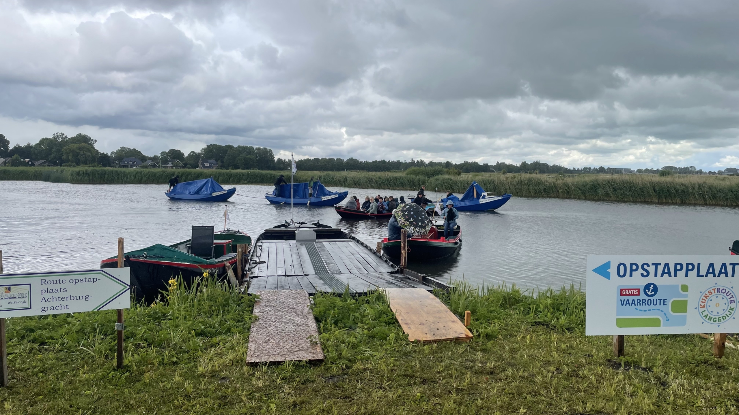 Een aanlegsteiger aan een rivier met meerdere boten waarop mensen zitten onder een bewolkte hemel, omgeven door grasachtige oevers. Een bord met de tekst 'OPSTAPPPLAATS' en een routebeschrijving is zichtbaar op de voorgrond.