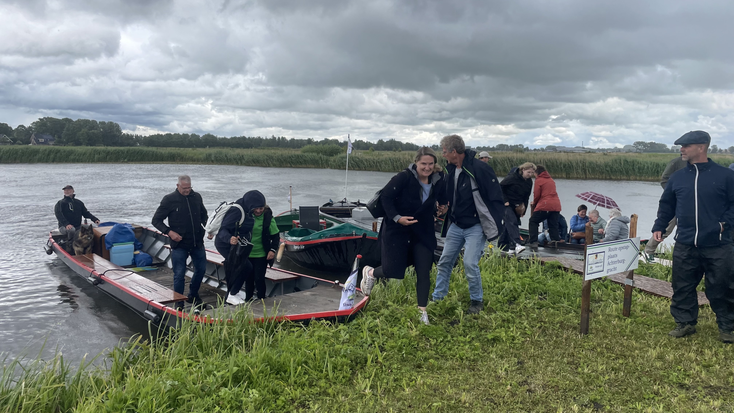 Mensen stappen uit boten aan de oever van een rivier onder een bewolkte hemel, omringd door groen gras en riet.