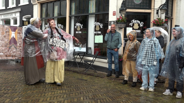 Straatoptreden met acteurs in historische kleding voor een bistro in een straat met vlaggen, terwijl toeschouwers in regenkleding toekijken.