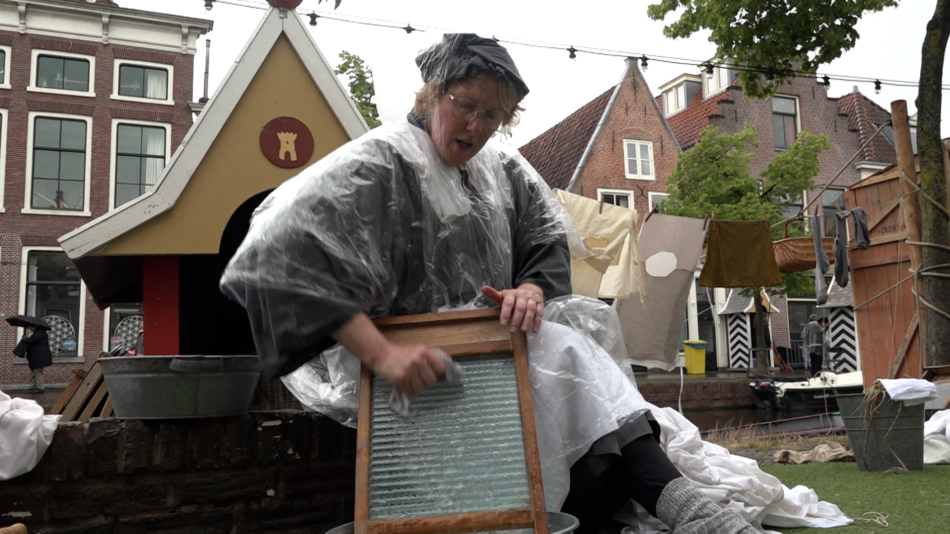 Een vrouw, gekleed in een plastic regenponcho, schrobt kleding op een wasbord bij een bak water in een historische setting, met op de achtergrond een waslijn en oude gebouwen.