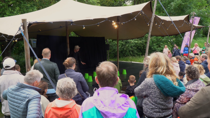 Groep mensen kijkt naar een optreden op een buitenpodium onder een tentdoek, omgeven door bomen.