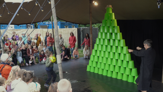 Een kind met een voetbal gooit naar een grote piramide van groene emmers, terwijl omstanders in een tent toekijken.