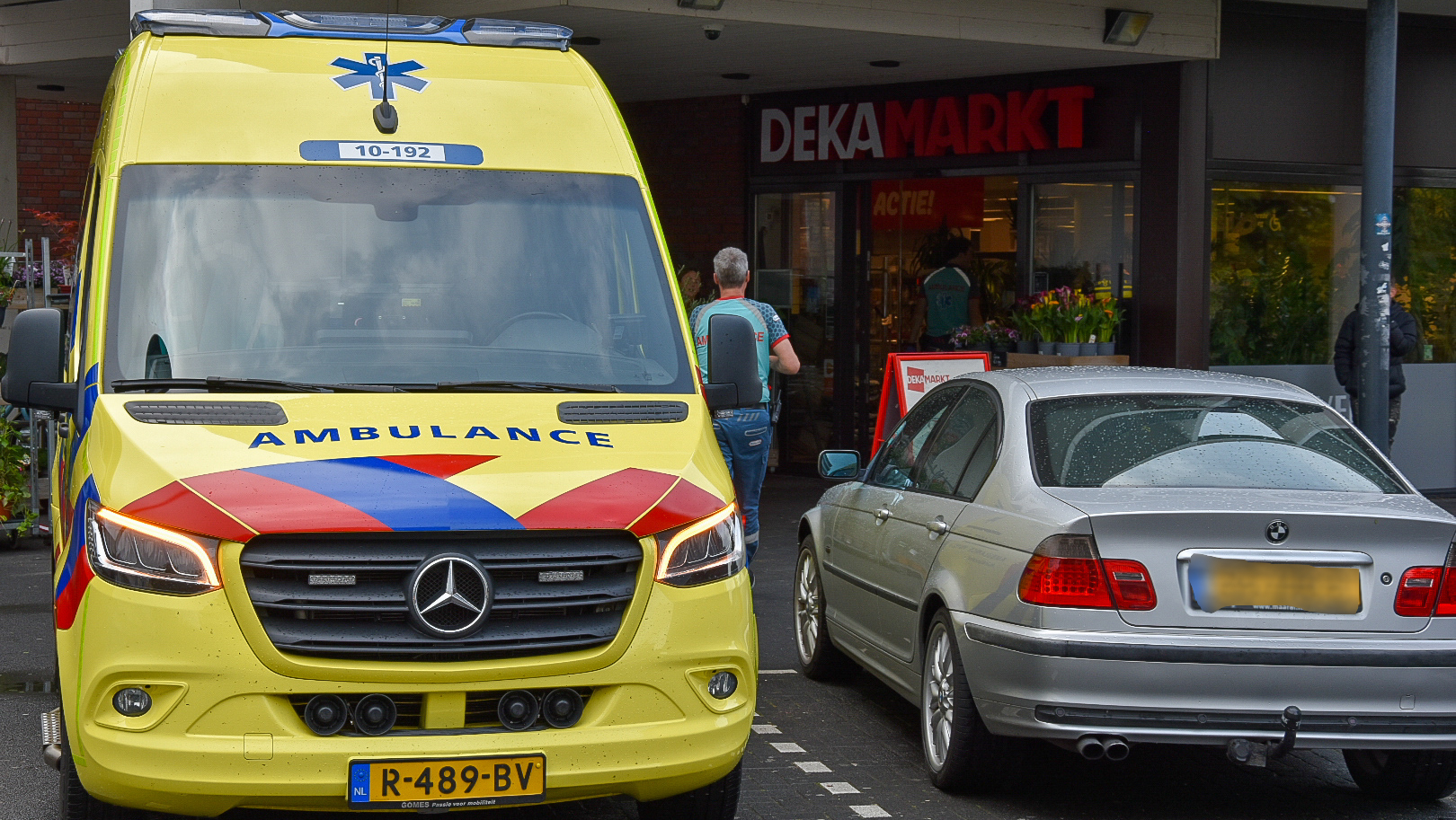 Een gele ambulance en een zilveren auto staan geparkeerd voor de ingang van een DekaMarkt supermarkt.