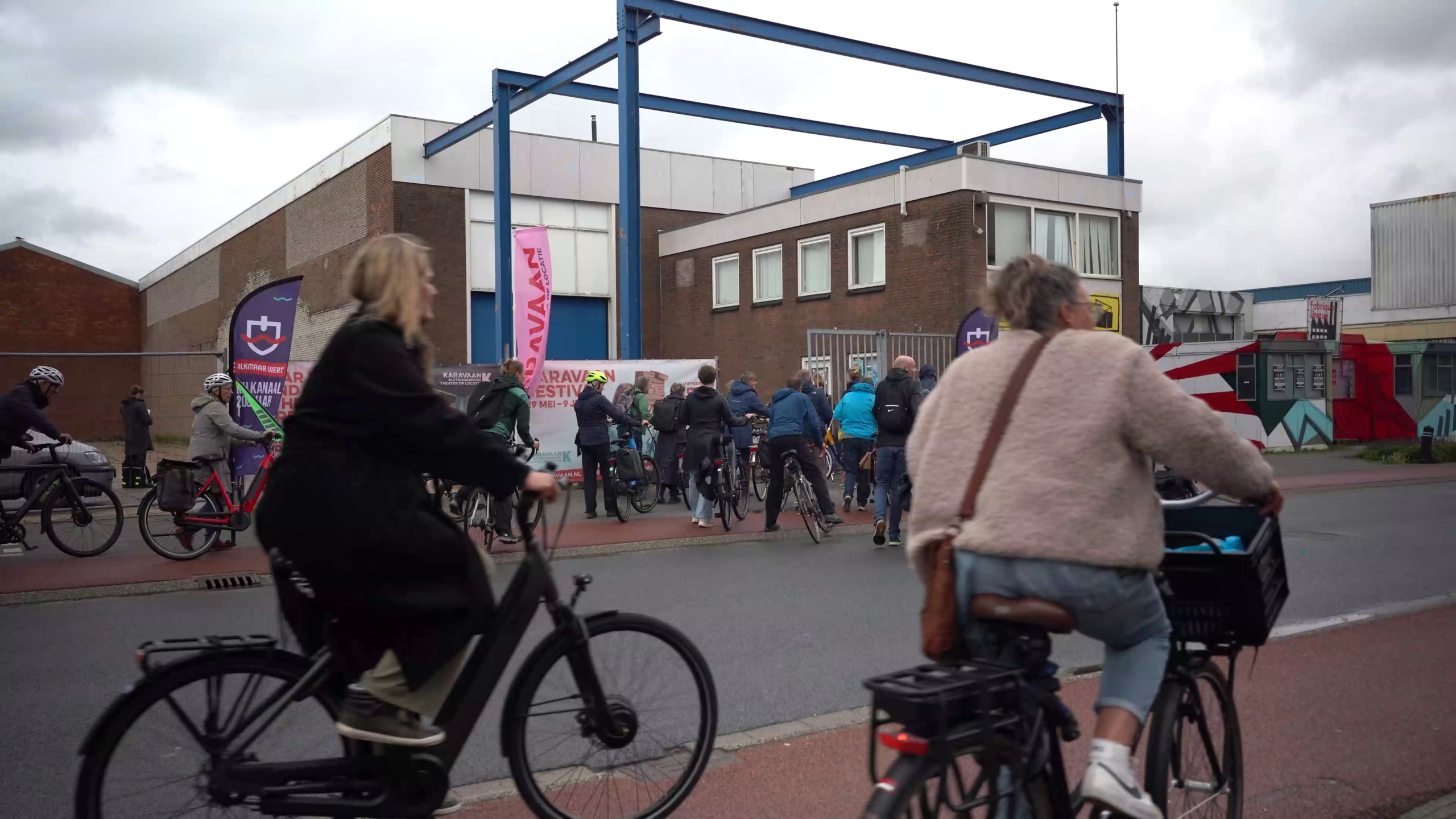 Mensen fietsen en wandelen bij een festivalterrein met bakstenen gebouwen en kleurrijke banners.