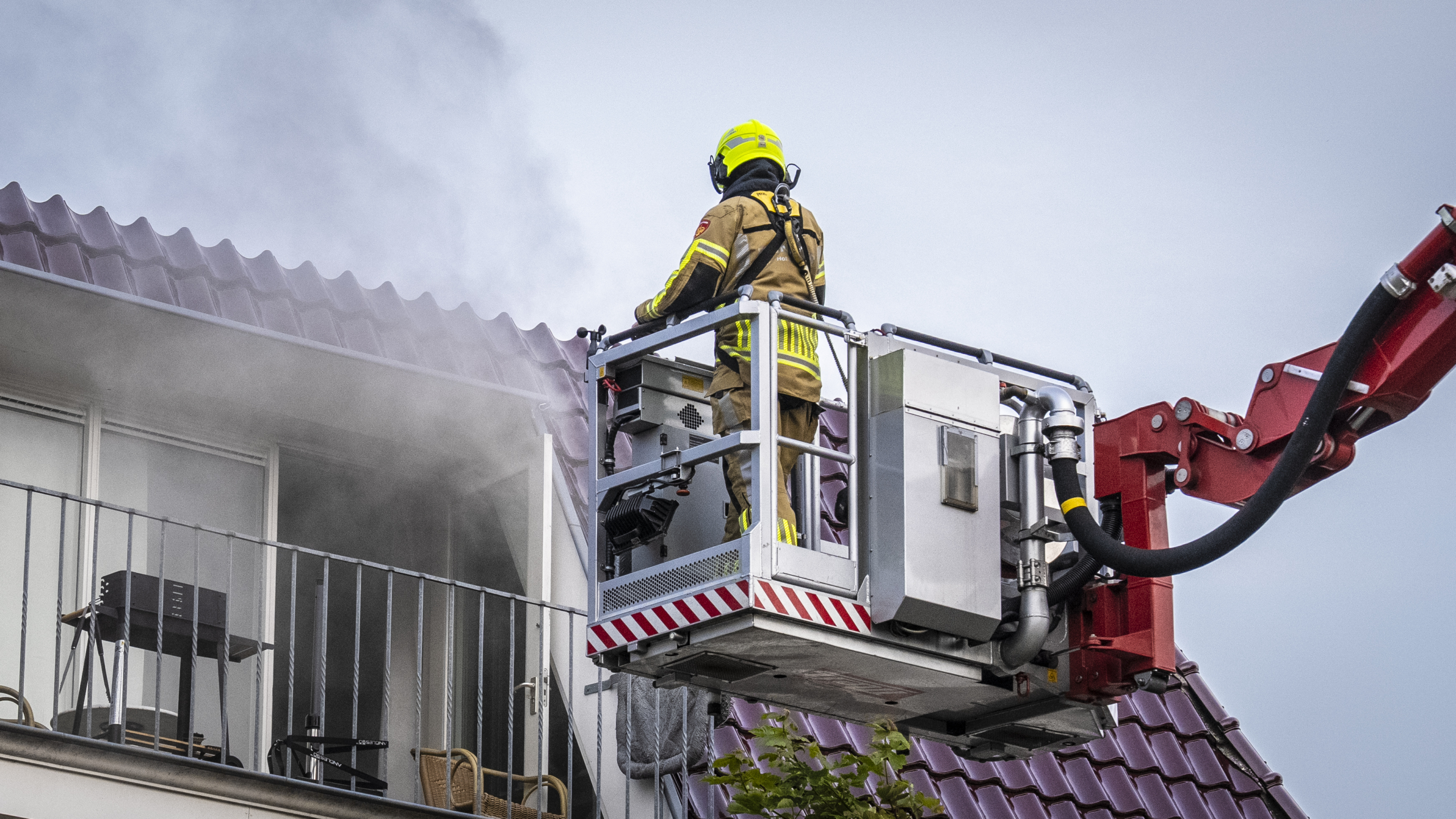 Een brandweerman staat in een hoogwerker naast een gebouw, terwijl hij rook bestrijdt die uit een raam komt.