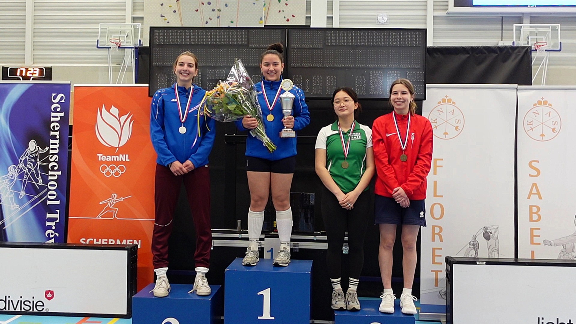 Vier vrouwen staan op een podium tijdens een schermevenement; de winnares in het midden houdt een beker en bloemen vast, terwijl de anderen medailles dragen. Op de achtergrond zijn schermbanners en een digitale klok te zien.