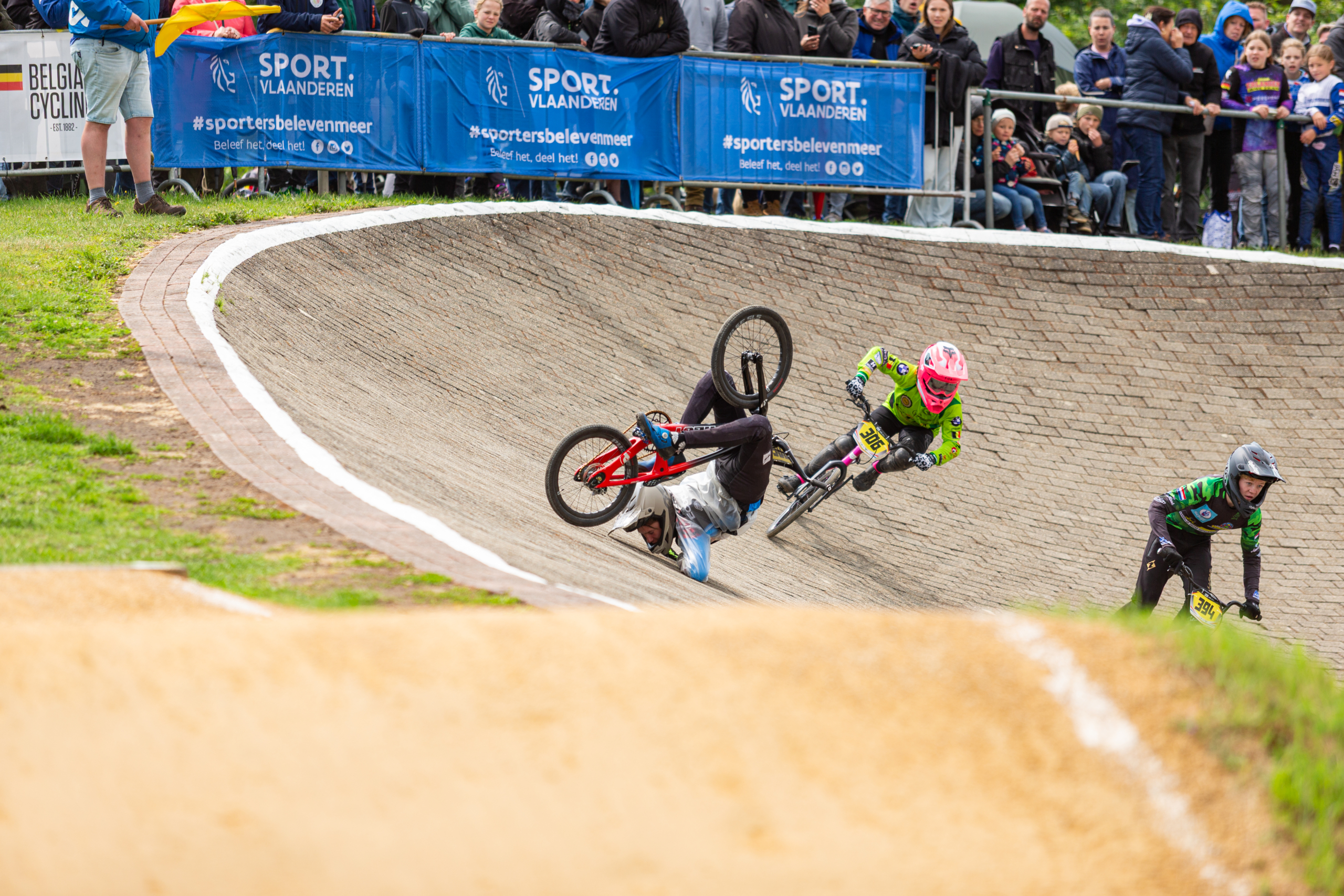 Een BMX-wedstrijd waarbij een fietser valt op een steile bocht, terwijl andere rijders in felgekleurde helmen verder racen voor een publiek langs de kant.