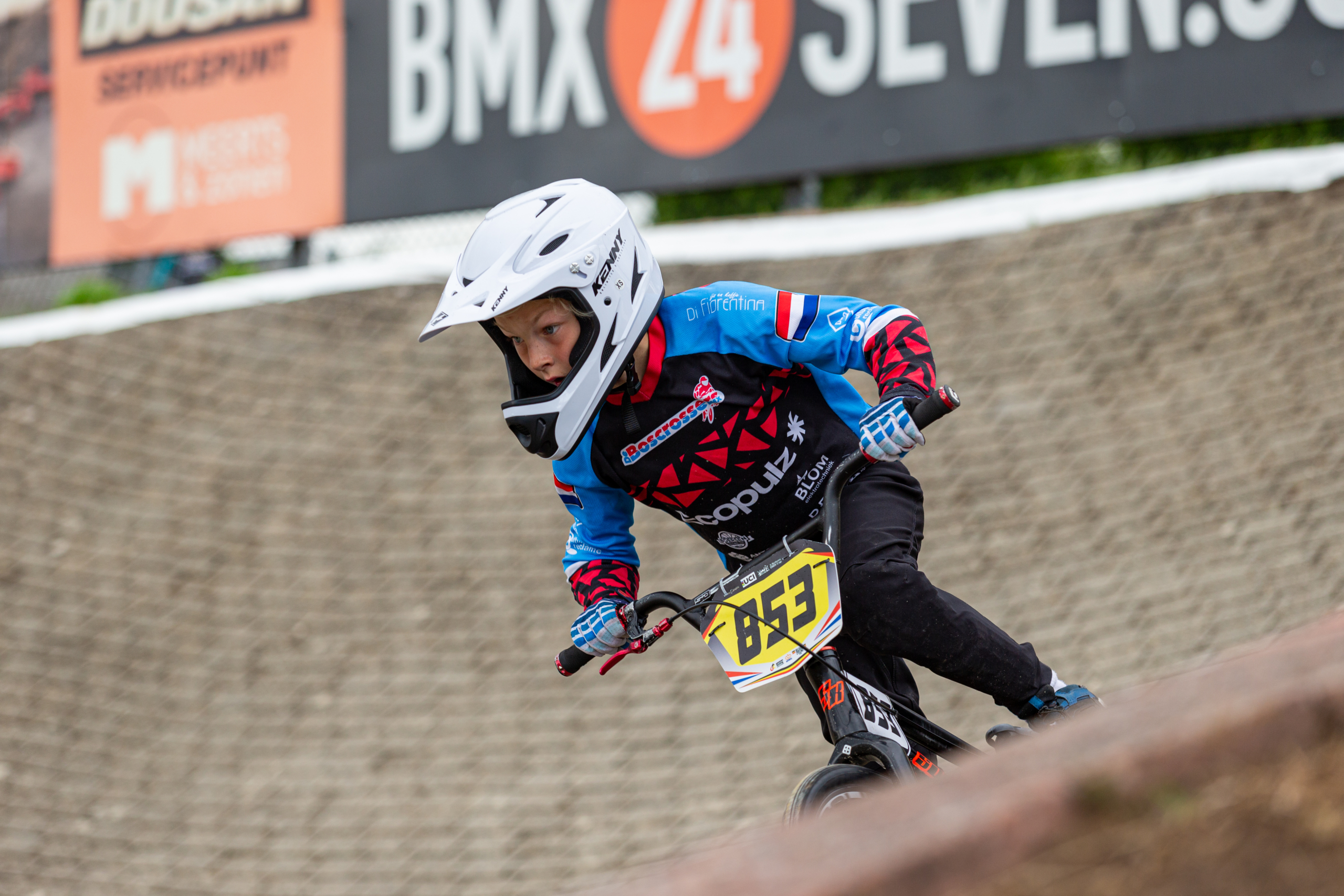 Een kind met een witte helm en blauw-rode BMX-outfit rijdt op een BMX-fiets met nummerbord 853 op een baanhelling, gefocust op de weg voor zich.