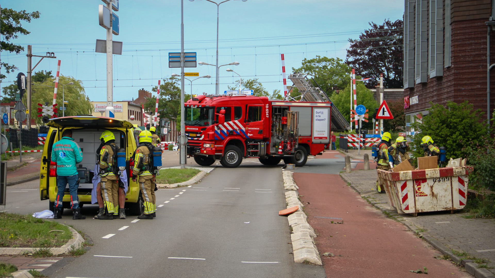 Brandweerlieden en een ambulancepersoneel staan naast een gele ambulance, terwijl een rode brandweerwagen de weg blokkeert bij een spoorwegovergang. Andere brandweerlieden werken bij een container aan de rechterkant van de weg.