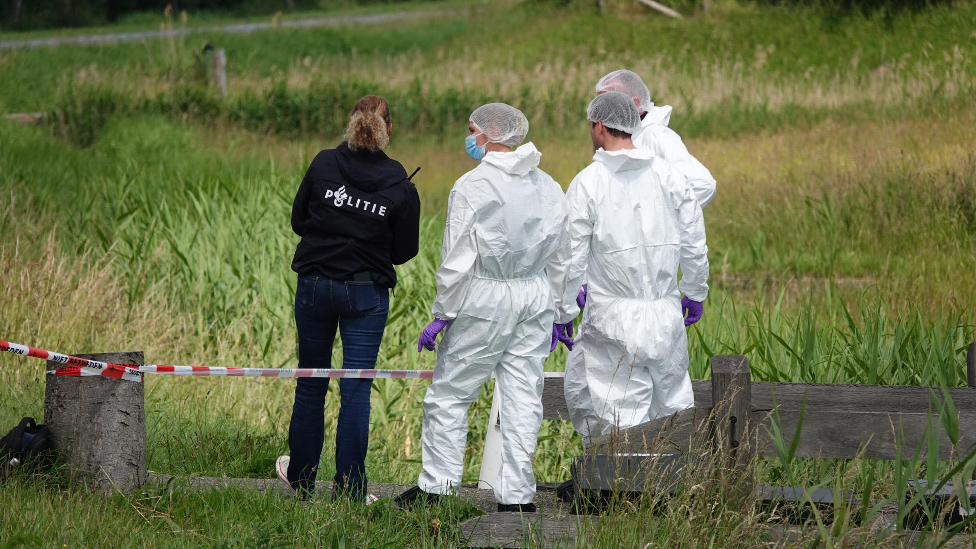 Een politieagent in een zwart jack en drie forensisch onderzoekers in witte beschermende pakken staan op een grasrijk gebied met een rood-witte afzetlint.