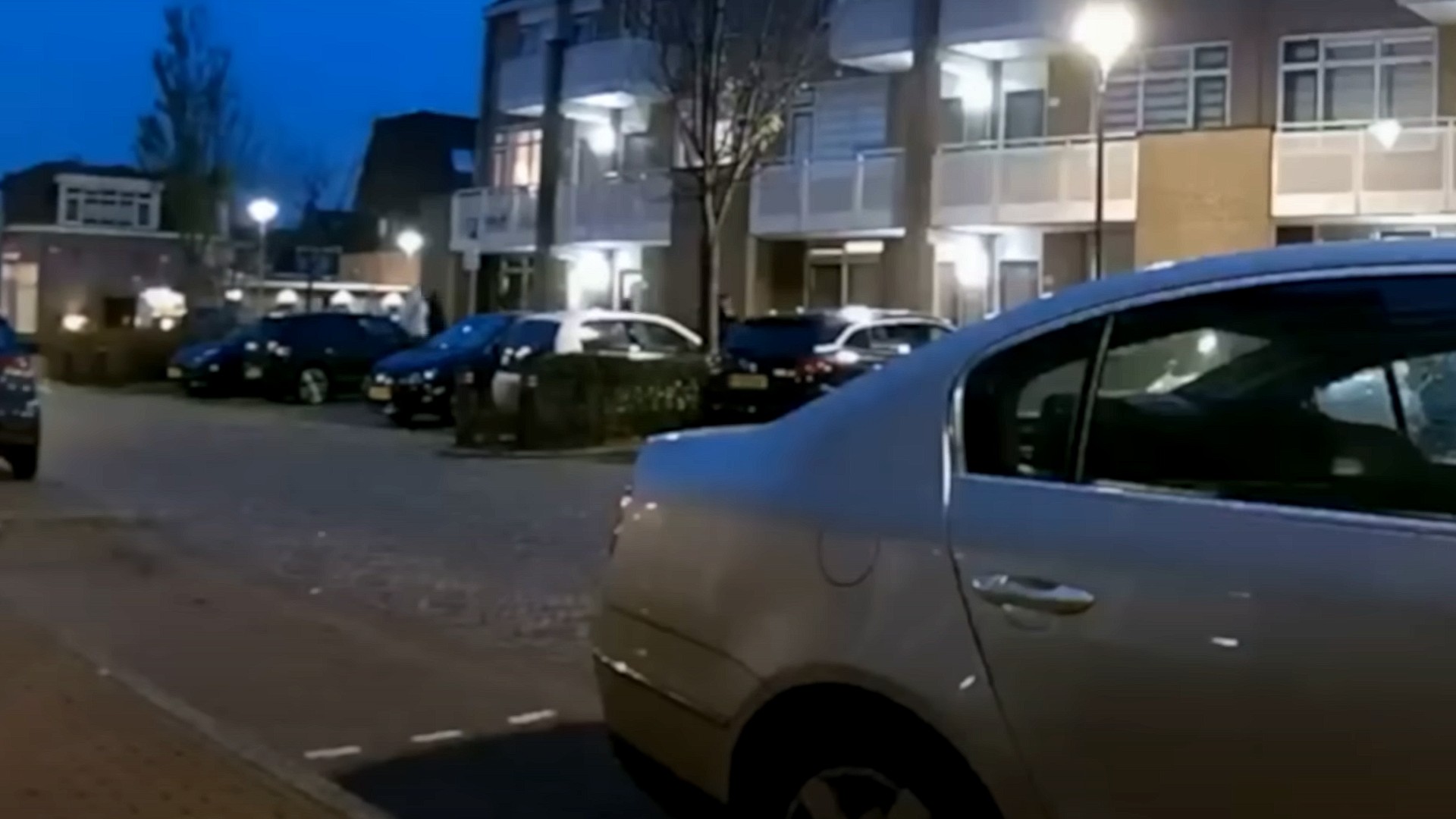 Straatbeeld in de schemering met geparkeerde auto's langs een rij huizen met verlichting.