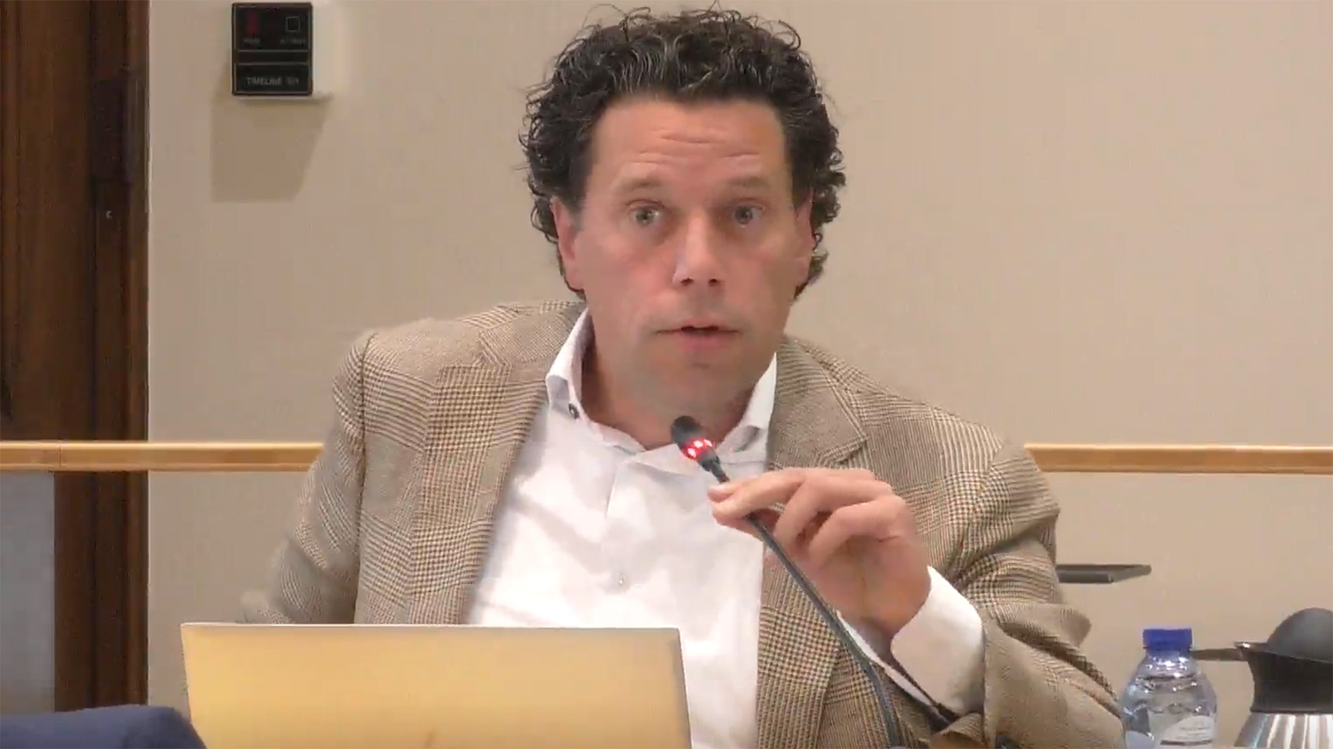 Man met een beige colbert en wit overhemd spreekt in een microfoon tijdens een vergadering, zittend achter een laptop.