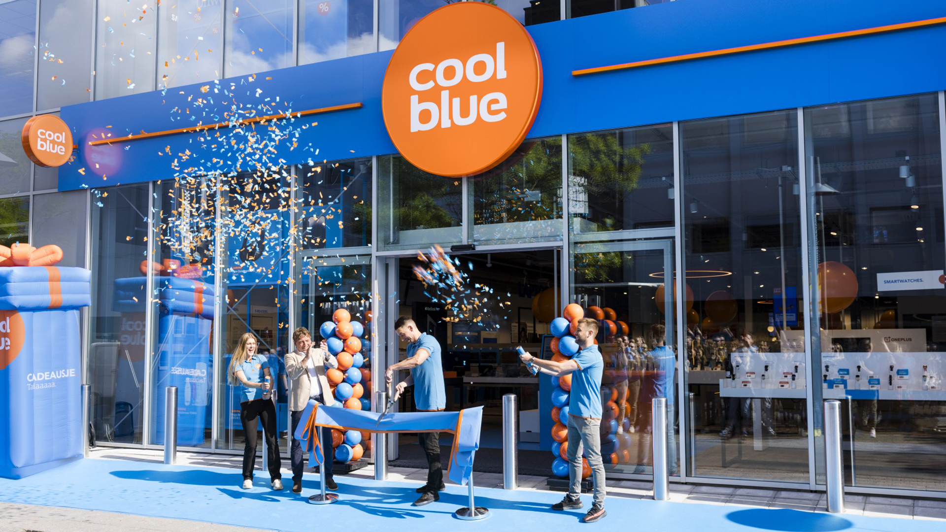Feestelijke opening van een Coolblue-winkel met mensen die confetti in de lucht schieten voor de ingang, versierd met blauwe en oranje ballonnen.