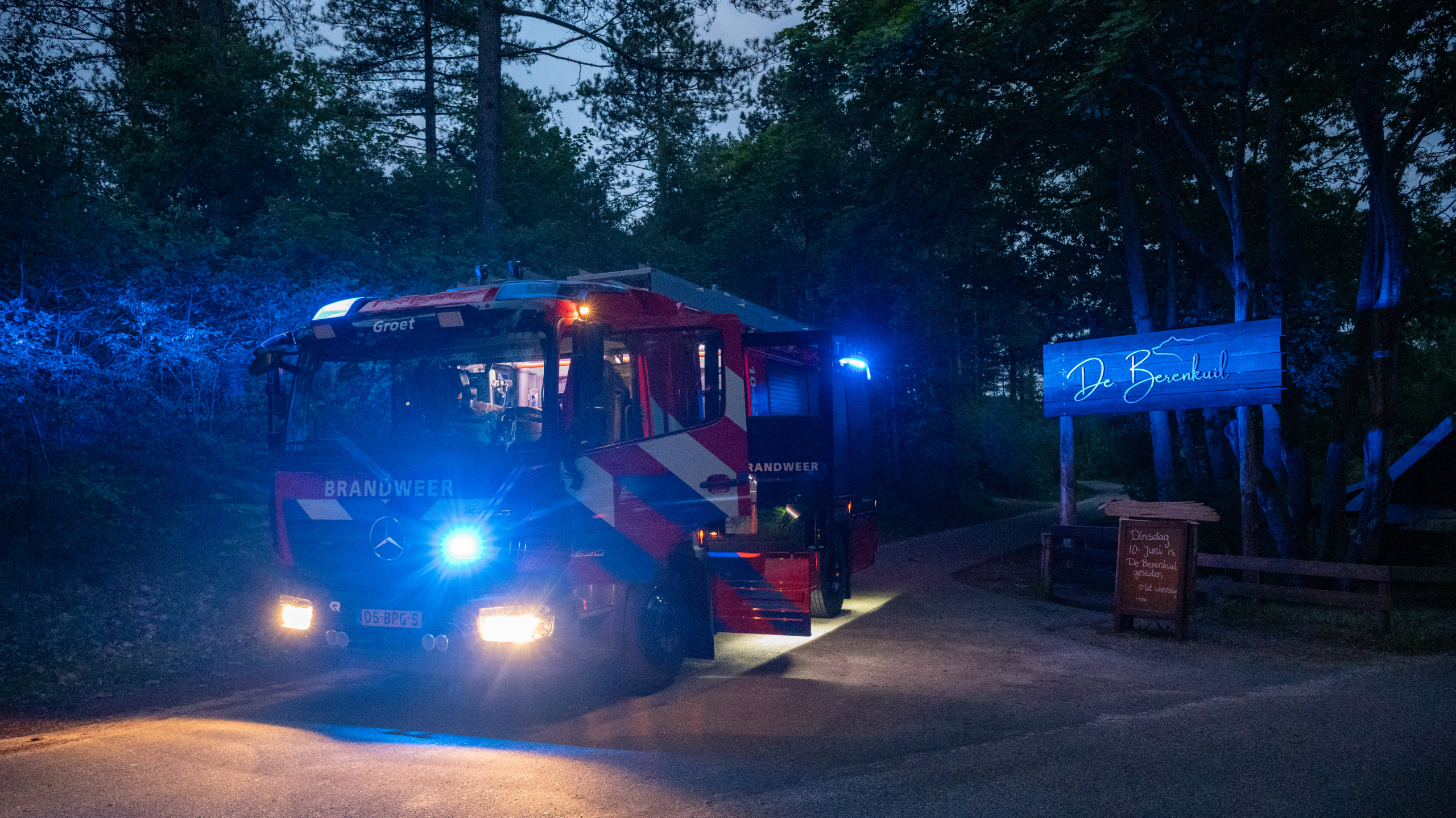 Een brandweerwagen met blauwe lichten aan, geparkeerd bij de ingang van De Berenkuil in een bosrijke omgeving, 's nachts.
