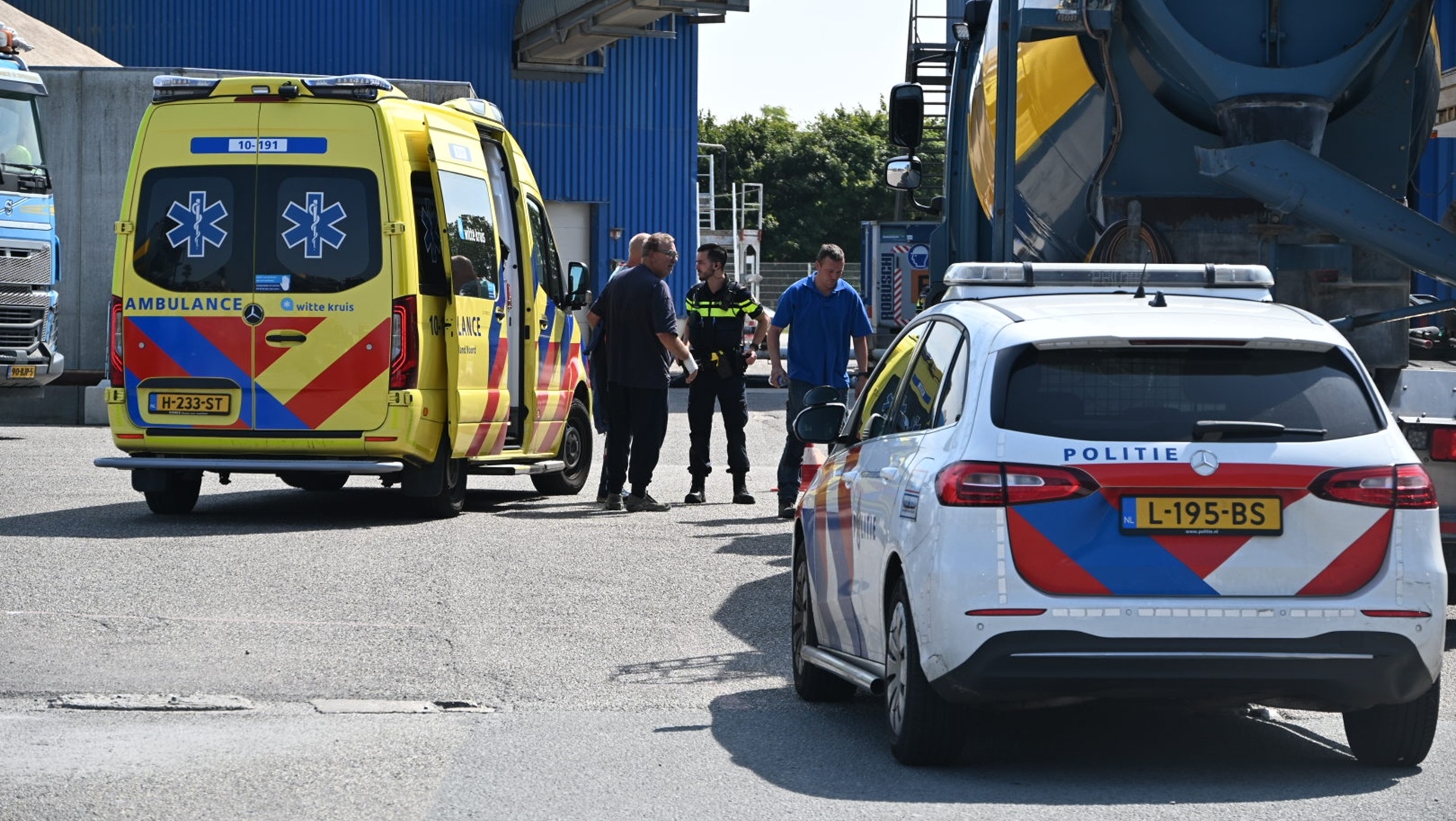 Een ambulance en een politieauto staan geparkeerd op een industrieterrein naast een blauwe vrachtwagen, met enkele personen eromheen, waaronder een politieagent in uniform.