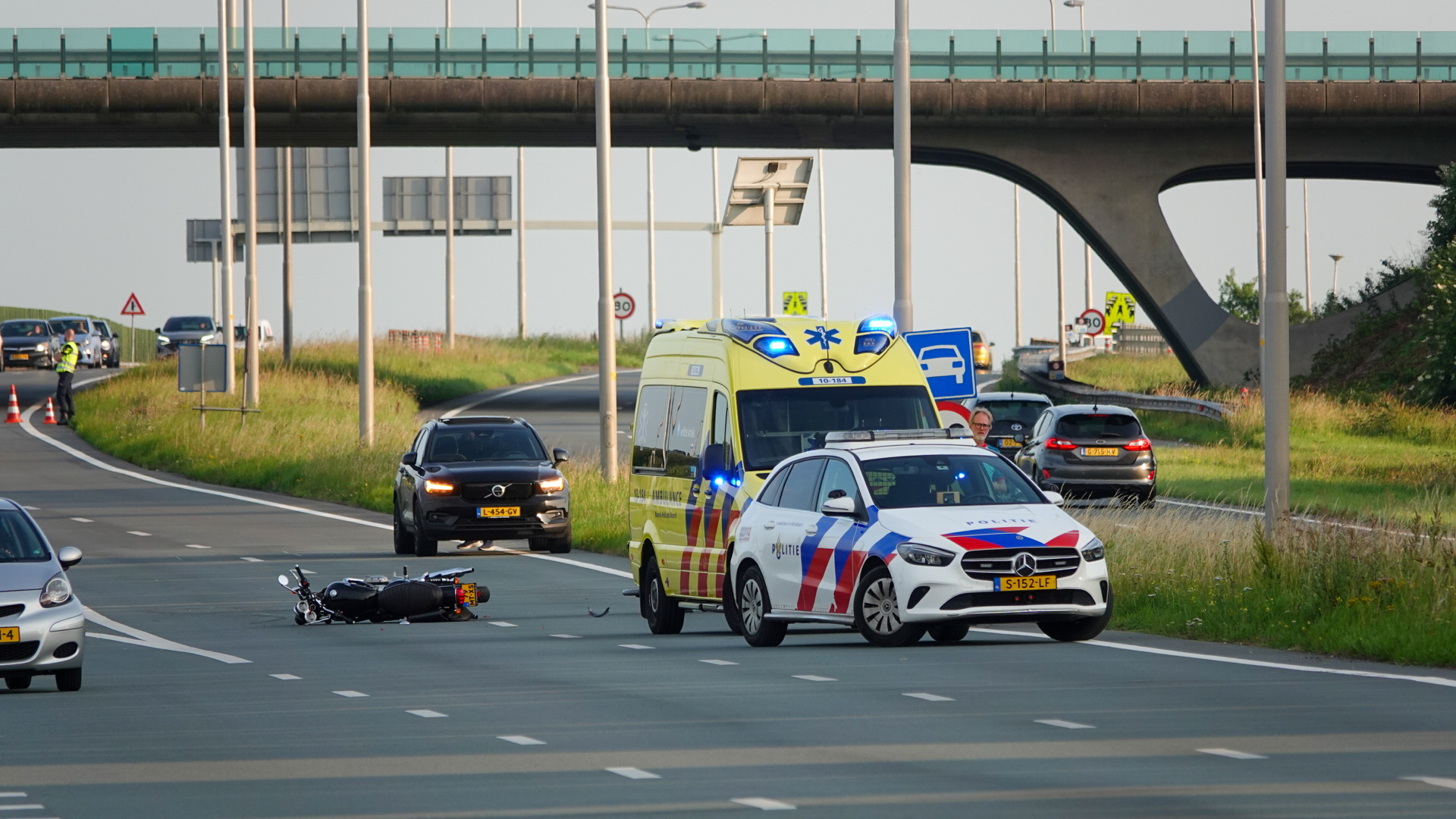 Ongeluk op de snelweg met een omgevallen scooter, omringd door een politieauto, een ambulance en een politieagent die het verkeer regelt.